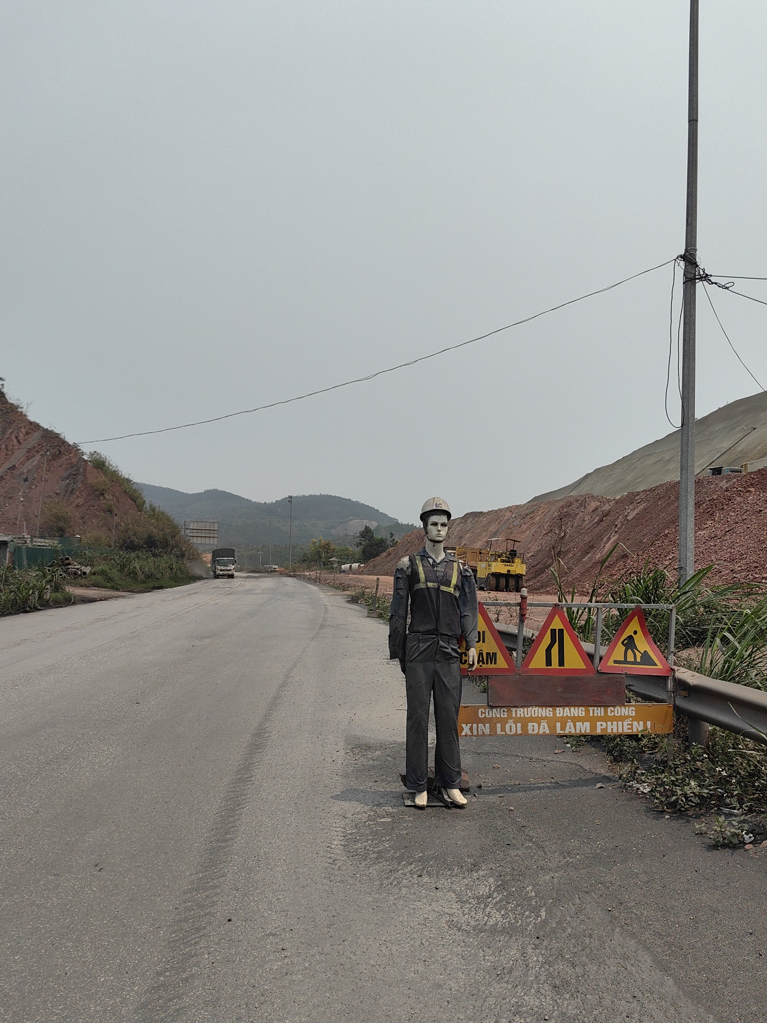 The image shows a roadside scene in what appears to be Thong Nhat Commune, Vietnam, evidenced by signage. A gray, paved road curves into the distance. A large, partially-constructed embankment made of red earth is visible on the right. There is a truck on the road in the distance.
In the foreground, a realistic mannequin dressed in construction worker attire stands beside a road barrier. The mannequin is wearing a hard hat, gray overalls, and has a blank expression. The road barrier features Vietnamese signage and triangular warning signs, including one with a figure digging. The barrier is positioned to block access to a section of the road. A utility pole is visible behind the barrier. Vegetation grows nearby.