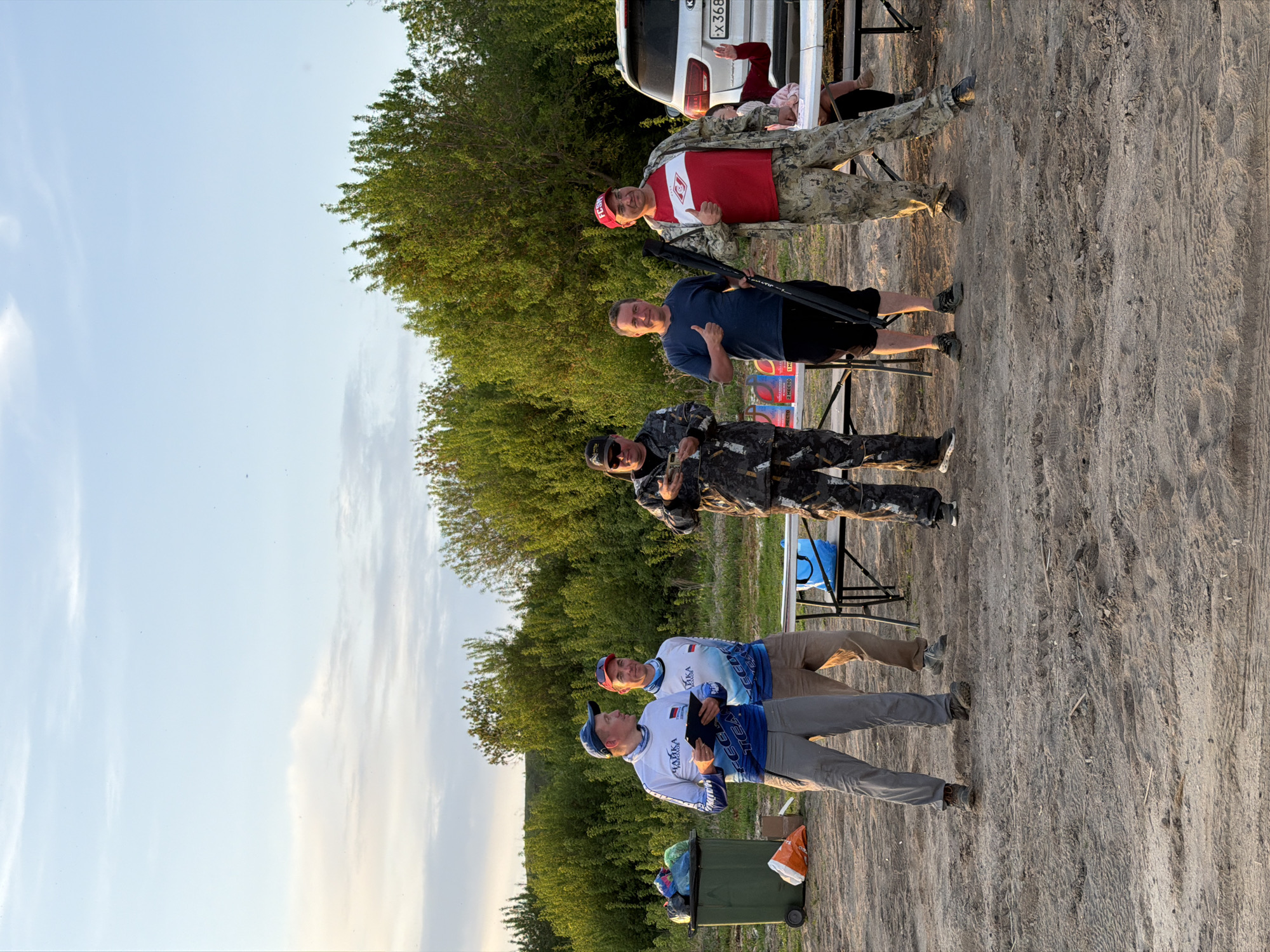 The image shows a group of six people standing outdoors, likely in the city of Krasnyi Bor, Russia, given that one of the team jerseys has the Russian flag on it. The ground appears to be sandy or dirt-covered. The weather is clear. In the background, there are trees and the rear of a white vehicle, where there's a person seated.
Five of the people are standing. Two men wear matching team shirts with the Russian flag on the sleeves. One man is dressed in camouflage clothing and is holding a small object, while another wears a blue shirt and stands with a black object, possibly a firearm. The last standing man wears camouflage pants and a red shirt with a white diamond.
At the rear of the vehicle is a person seated.