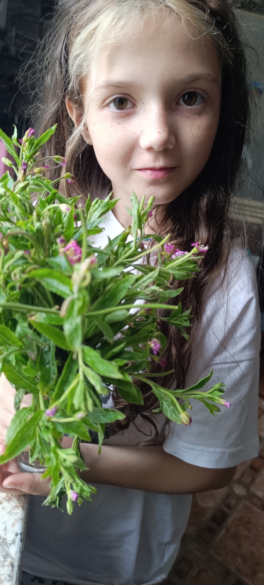 The image shows a close-up of a young girl with brown and blonde hair, freckles, and brown eyes, holding a bouquet of green leafy plants with small purple flowers. She is wearing a white t-shirt. The background is out of focus and appears to be an indoor setting with tiled flooring visible at the bottom right. The girl's face is partially obscured by the plants. There is no indication of the city of Karsha, Russia, in this image.