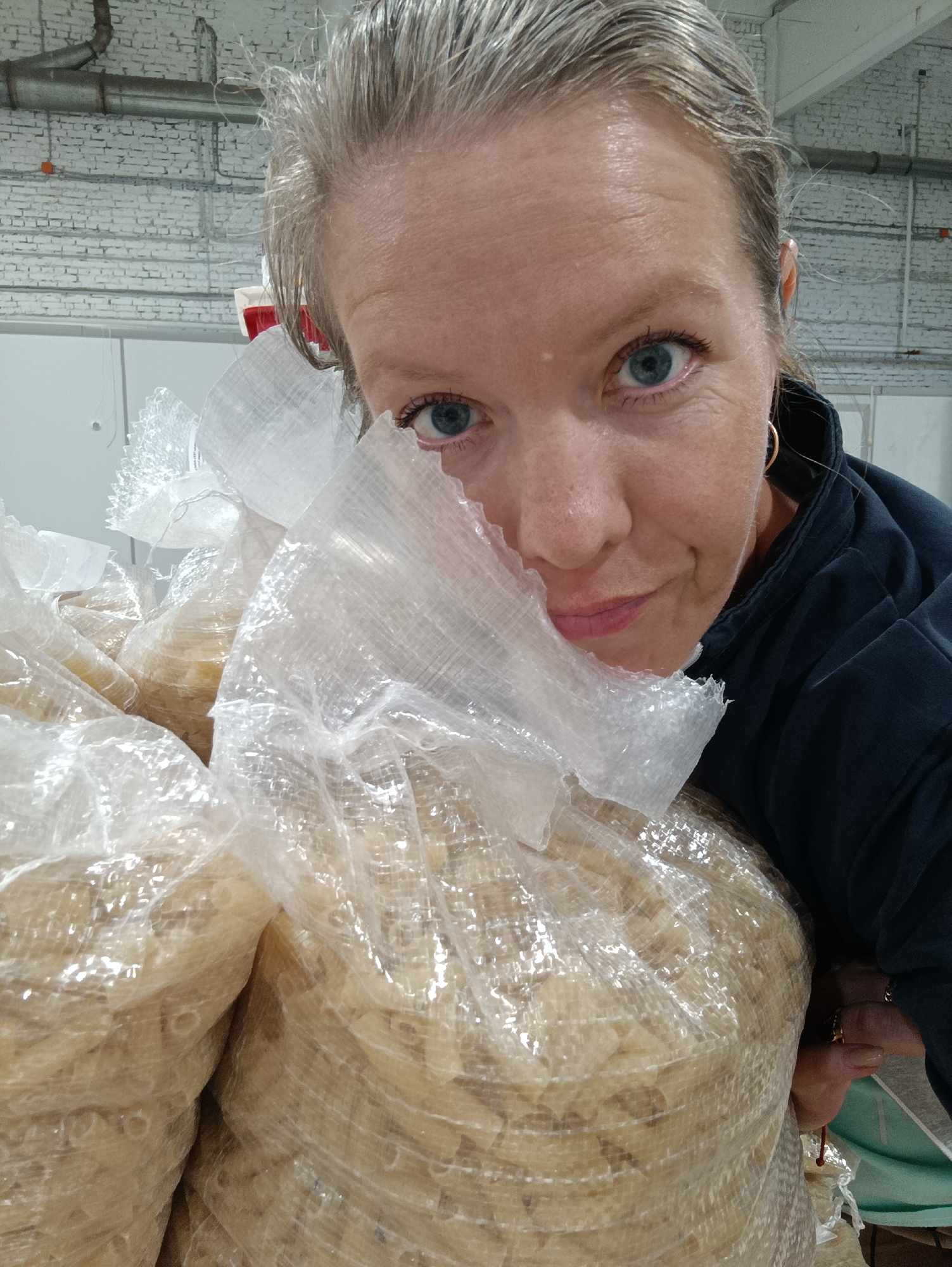 A woman is posing close to several large bags filled with what appears to be dried pasta or a similar food product. The bags are made of clear plastic. The woman has light skin, graying hair pulled back, blue eyes, and is wearing hoop earrings and a dark blue or black top. Her right hand is visible holding one of the bags. The background includes an industrial-looking area with white brick walls, ceiling pipes, and possibly partitioned areas. The location is Chaikovsky, Russia.