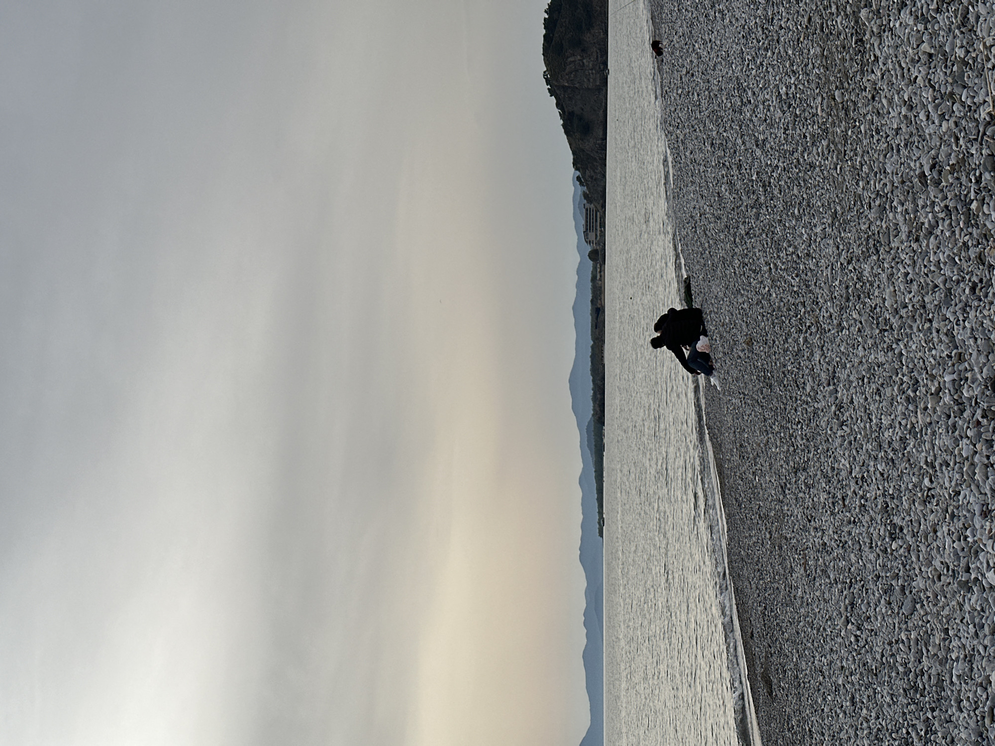 The image shows a coastline in Fethiye, Turkey. The sky is overcast, casting a soft, diffused light. To the right of the frame, a pebble beach slopes down to the water's edge. A person is crouched near the shoreline, possibly interacting with something on the ground. In the background, a mountainous terrain rises from the horizon, with a few buildings visible along the slope. The water appears calm with gentle ripples.