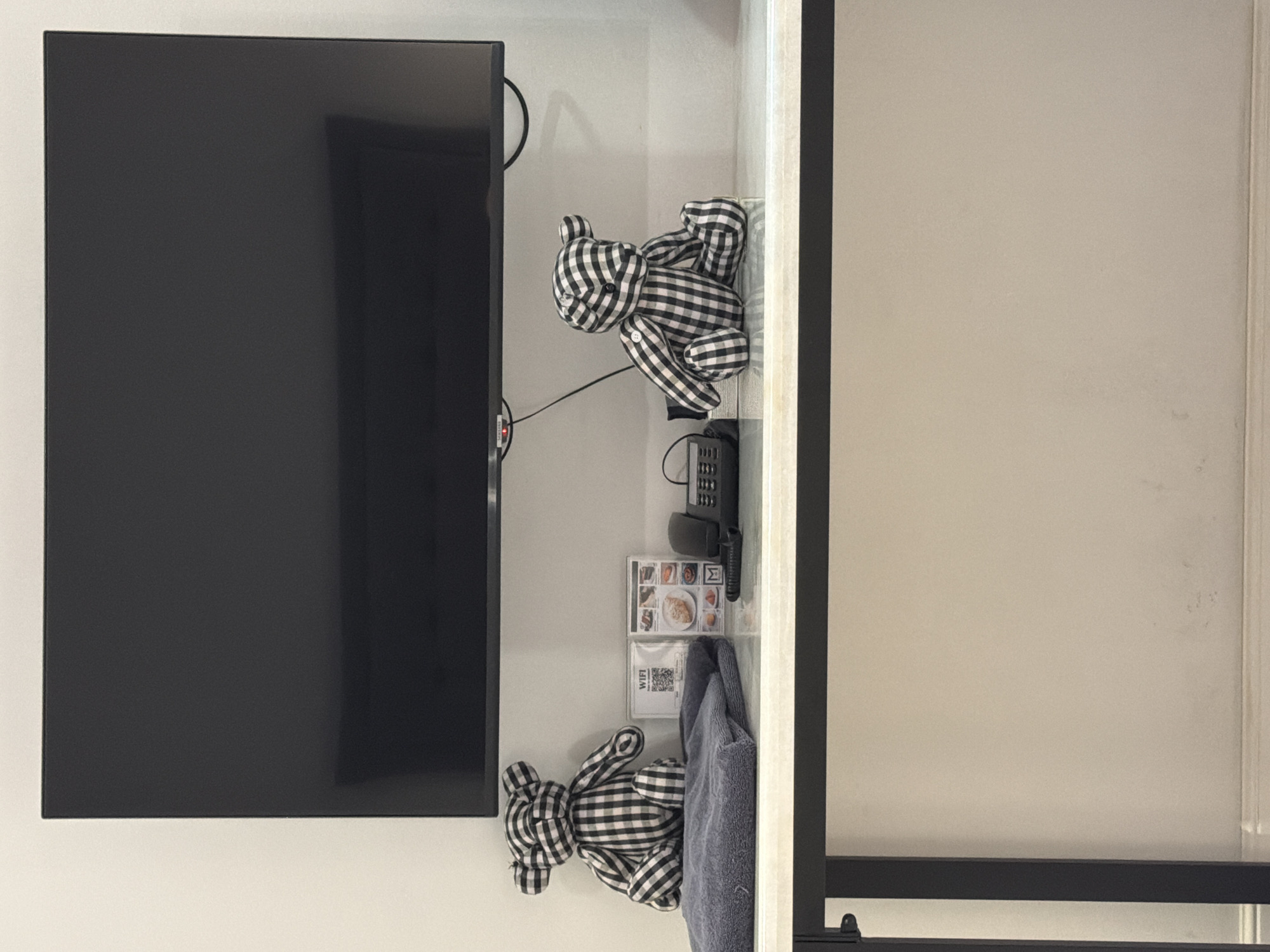 A black and white checkered teddy bear sits on a stack of folded gray towels on a marble-topped table. To the right of the towels, a small framed sign with a QR code and "WIFI" printed on it stands next to a framed picture menu. Further to the right, a black telephone with a coiled cord is positioned next to another checkered teddy bear, this one also sitting. Above the table, a large black flat-screen television is mounted on a white wall. The scene appears to be set in a hotel room in Ban Phonsavang Neua, Laos.