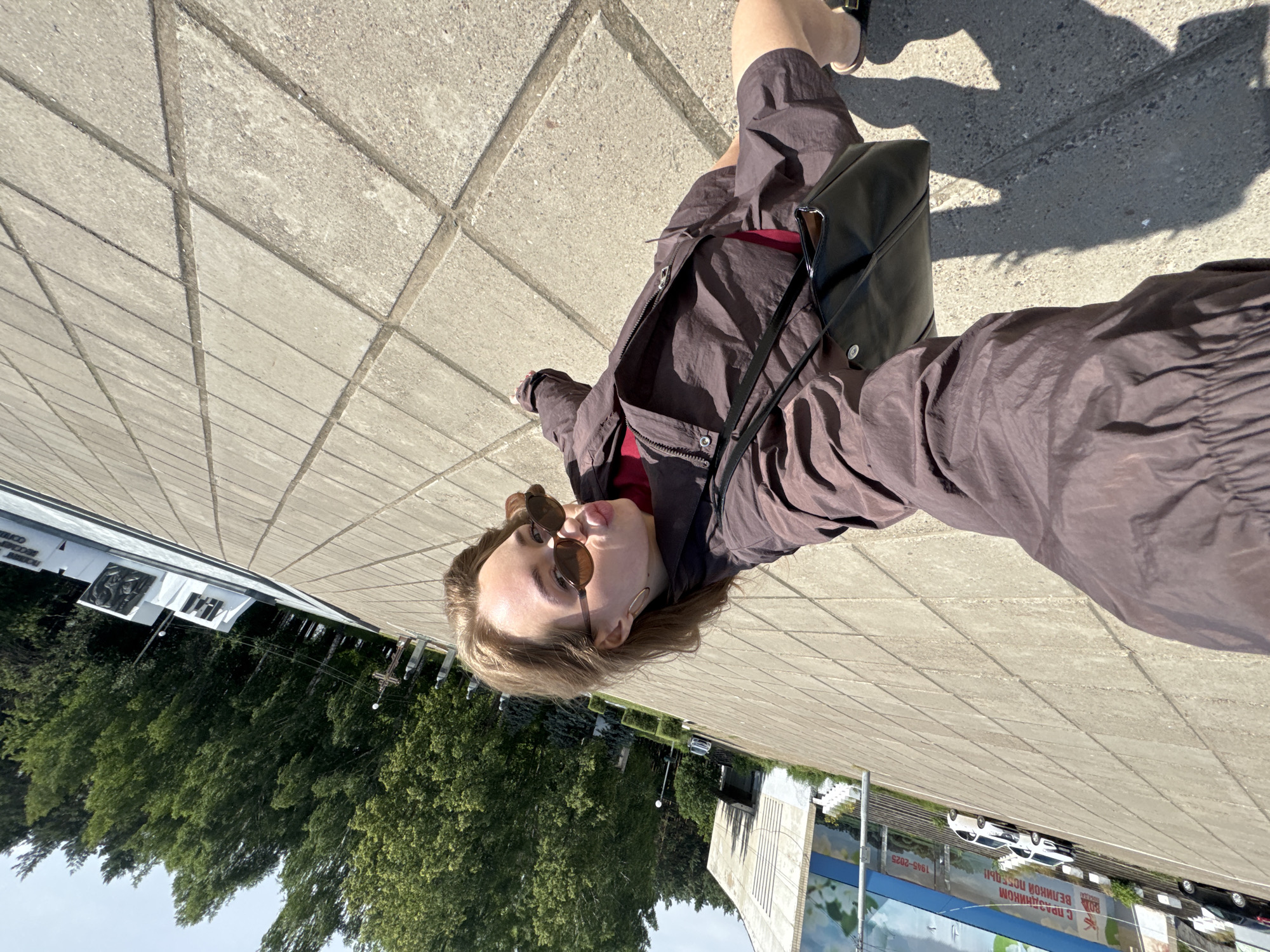 A young woman with blonde hair and sunglasses is taking a selfie outdoors in Chaikovsky, Russia. She is wearing a brown jacket and shorts with a red shirt underneath. A black bag is slung over her shoulder. The ground is paved with concrete tiles, and in the background, there are trees, a building, and a monument. The sun is shining, casting shadows on the ground.