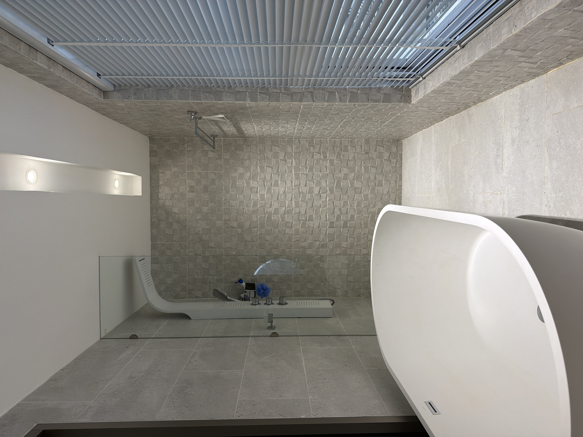 This is a forensic, matter-of-fact description of a modern bathroom, likely located in Thep Krasatti, Thailand. The scene depicts a freestanding white bathtub positioned in the foreground, taking up the left half of the frame. To the right of the bathtub is a shower enclosure with a clear glass door. Inside the shower, a modern white shower panel with multiple controls and a showerhead is visible. A blue loofah hangs from the shower panel.
The bathroom walls are tiled with large, light gray rectangular tiles on the left side, transitioning to smaller, textured gray tiles on the back wall. A towel rack with a white towel hanging from it is mounted on the back wall. To the far right, a large window with white horizontal blinds is present. The floor is tiled with large, light gray square tiles. The ceiling is white with recessed lighting, including a linear fixture with two circular lights. The overall impression is one of a clean, contemporary, and well-maintained bathroom. There are no people or discernible actions taking place.