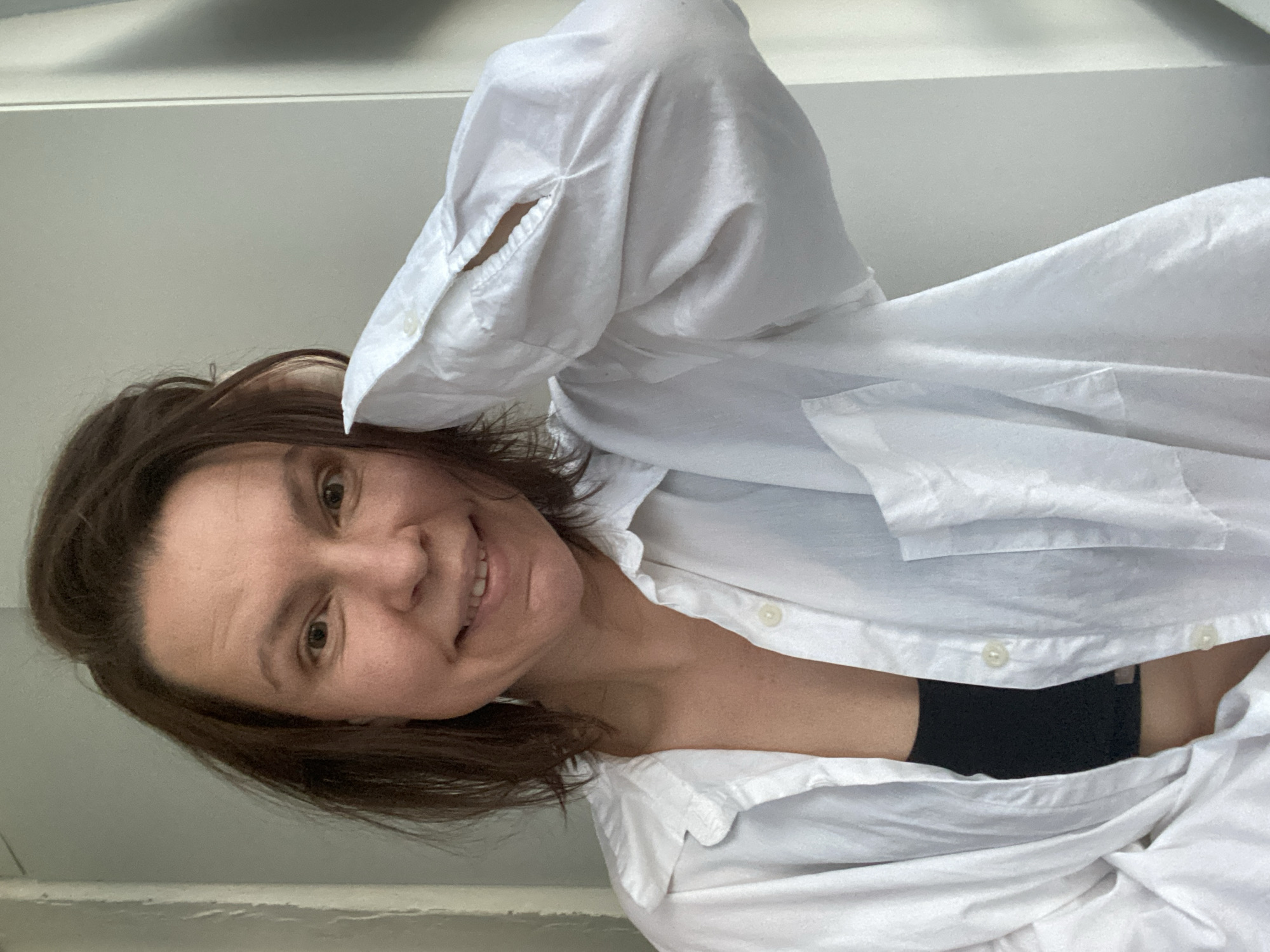 A woman with brown hair is wearing a white button-down shirt and a black undergarment. The shirt is partially unbuttoned and draped around her. The background appears to be a light-colored wall. No specific location in the city of Chaikovsky, Russia, can be identified from the image.