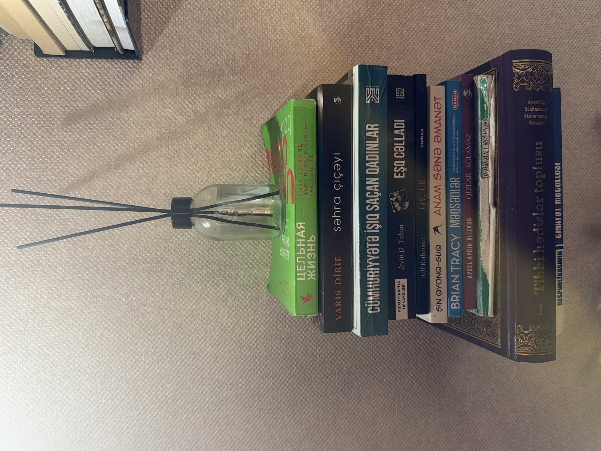 A stack of books is placed on a shelf against a textured, beige wall. A glass bottle with reeds, likely an air freshener, stands on top of the books. The books are varied in size and color. The top book is bright green with text in Russian. Below that are dark-covered books with titles in a foreign language, some appearing to be in Cyrillic script and others in Latin script. The bottom-most book is large and dark purple with ornate gold lettering. A part of a second stack of books is visible to the right of the main stack, with their spines facing outwards. The lighting suggests an indoor environment, possibly a home or office. There are no people visible in the image. The image does not contain any information that would allow for determination of the location within Sabirabad, Azerbaijan.