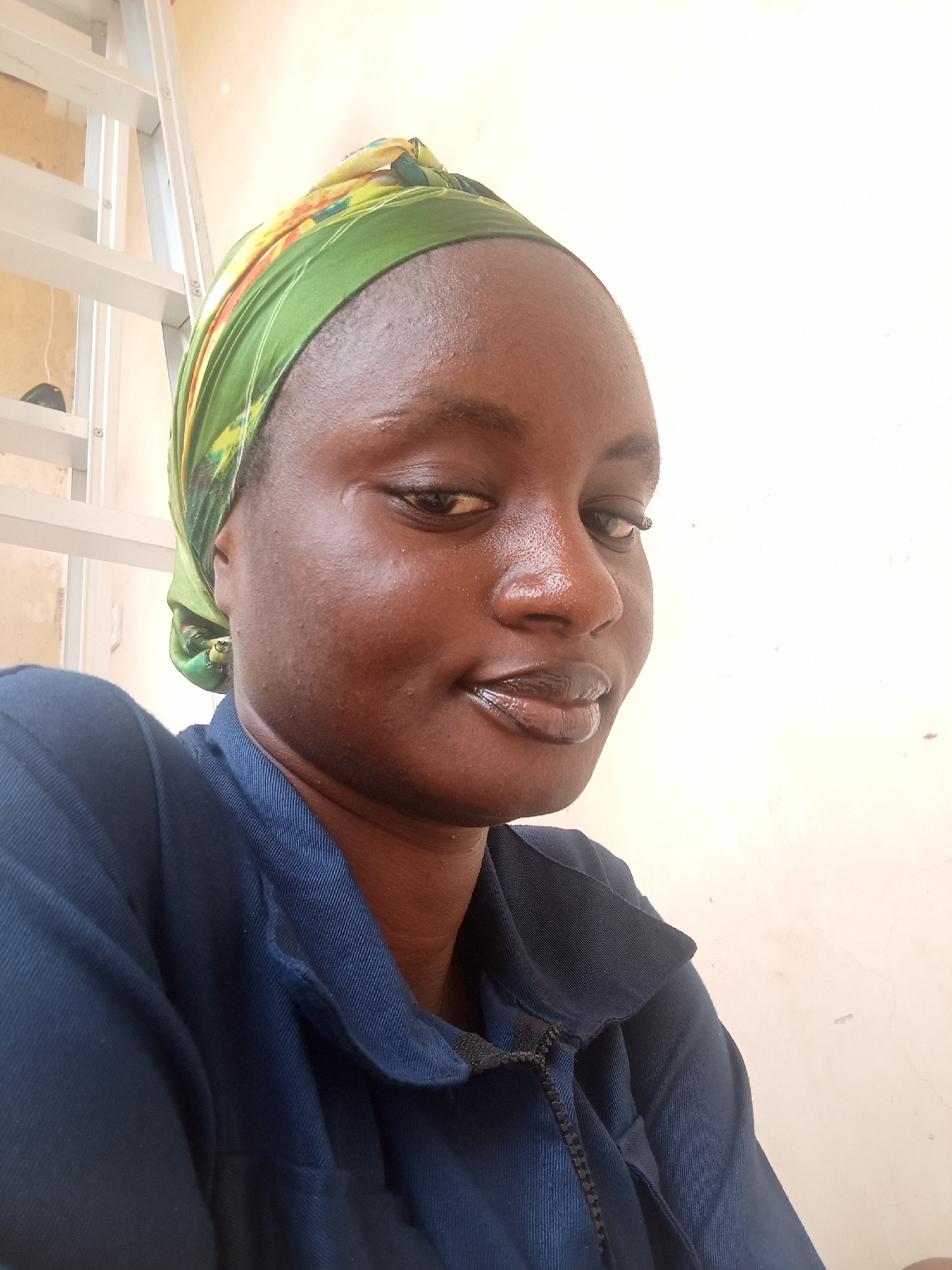 A close-up, medium-angle shot shows a young woman with dark skin and a green and yellow headscarf framing her face. She is wearing a dark blue collared shirt with a zipper. Her head is slightly tilted to the left, and she is looking directly at the camera with a slight smile. Her eyes are dark brown, and her lips are glossy. The background is a light-colored wall with a white ladder to the left of the woman. The overall lighting is bright and appears to be natural daylight. The image was taken in Shiroro, Nigeria.