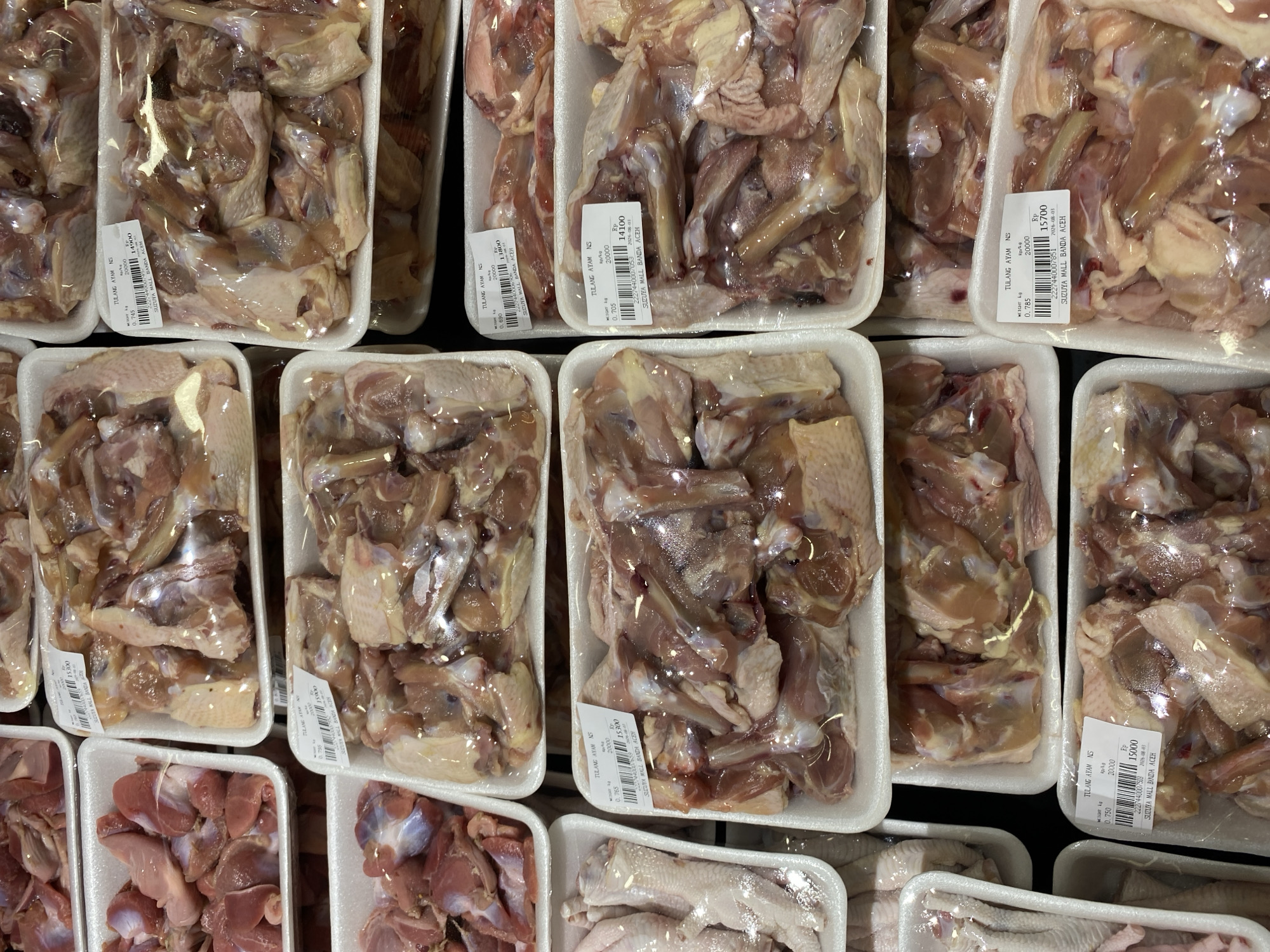 The image shows a top-down view of several plastic trays filled with raw chicken pieces, arranged in a retail display. The chicken pieces vary in size and cut, with some appearing to be bone-in portions and others possibly smaller cuts. Each tray is sealed with plastic wrap. Some of the trays have white labels with text in what appears to be Indonesian, including pricing and product information. One label clearly reads "SUZUYA MALL BANDA ACEH", indicating the location as a Suzuya Mall in Banda Aceh, Indonesia. The overall environment suggests a grocery store or supermarket meat section.