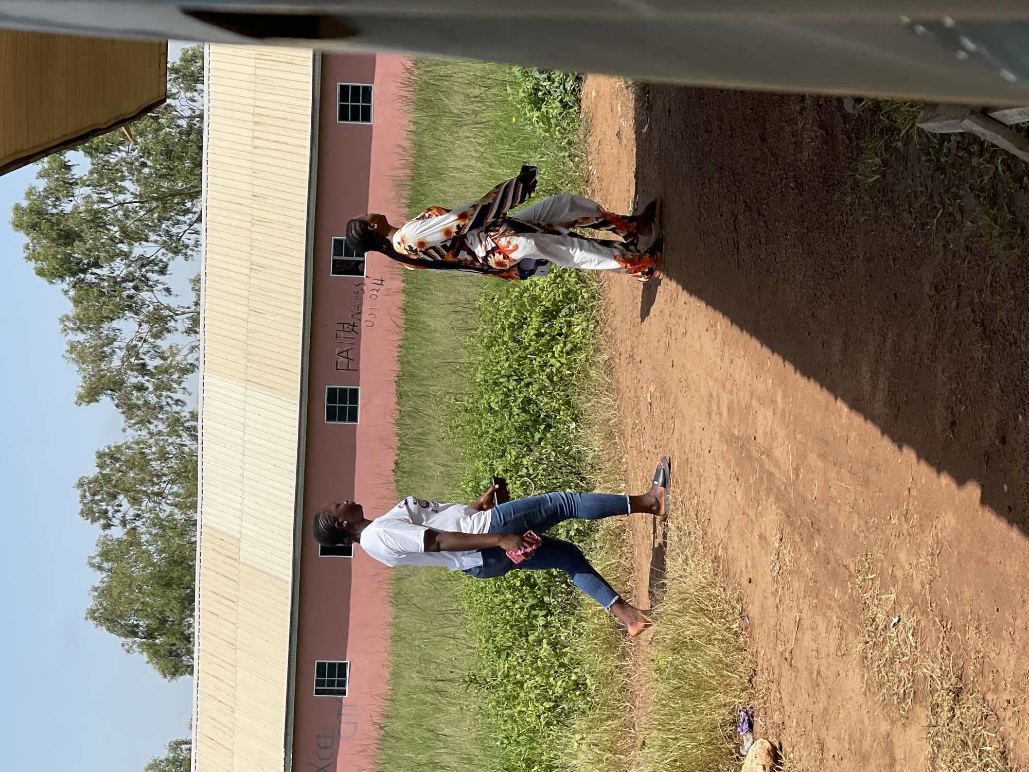 In Durumi, Nigeria, two women walk on a dirt path next to a pink building with a light-colored metal roof. The woman on the left wears a white t-shirt and blue jeans, carrying a phone and a pink case. The woman on the right is dressed in a colorful, patterned outfit with long sleeves and pants. The building has several windows and graffiti on its facade, including the words "FAITH NAPSS" and numbers. Green vegetation and trees are visible in the background. The sun casts shadows across the path.