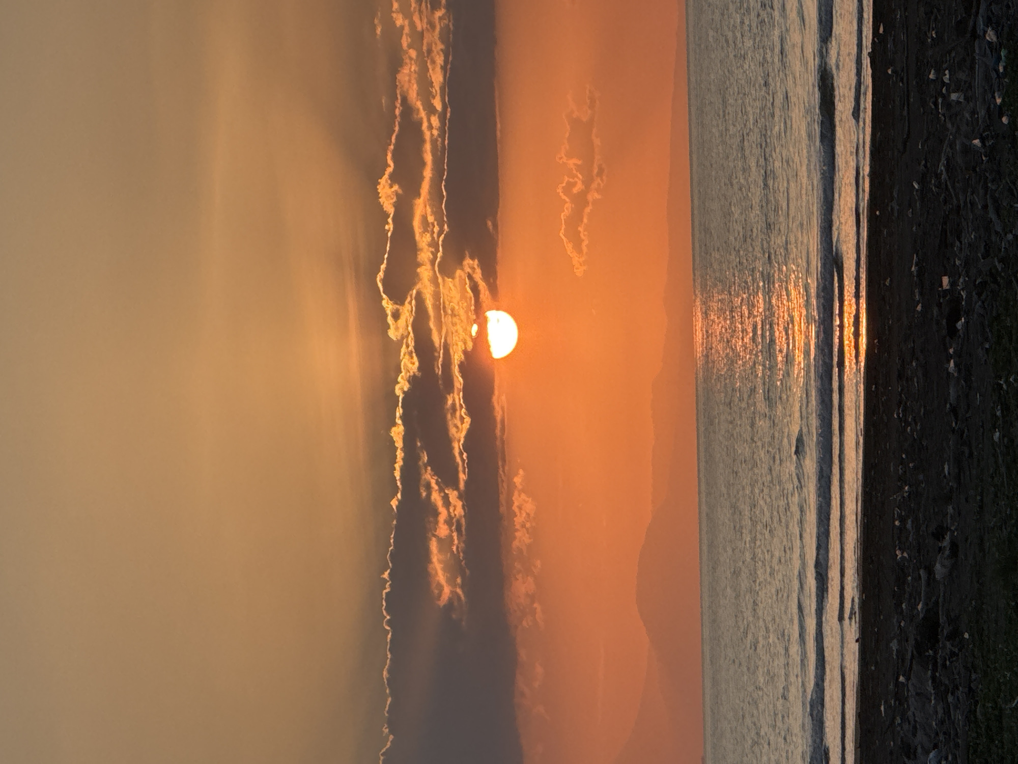 This is a vertical, medium-shot photograph of a sunset over the ocean, taken from the beach in Meunasah Jurong, Indonesia. The sky is a warm gradient of orange and yellow, with a large, partially obscured sun near the center of the frame. Dark, textured clouds are scattered across the sky, with the sun's light filtering through them. Below the sky, distant, hazy mountains are silhouetted against the horizon. The ocean is a calm, rippling body of water reflecting the sunset. Gentle waves are breaking on the shore. The foreground is a dark, sandy beach littered with debris, including plastic waste and other refuse, with patches of green vegetation at the bottom edge of the frame. No people or animals are visible in the scene. The overall impression is one of natural beauty juxtaposed with environmental pollution.