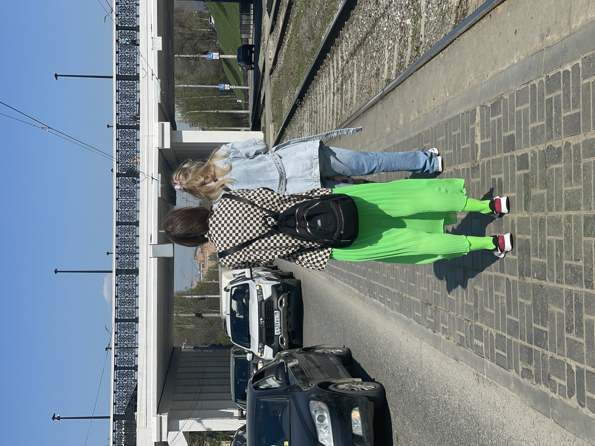 Two people are standing on the sidewalk of a road on the bridge. The first person has blonde hair, a denim jacket, and blue jeans. The second person has a black and white checkered jacket, a black backpack, and a bright green skirt with sneakers. There are cars traveling along the road in both directions on the bridge, and railway tracks are on the other side of the sidewalk. The white car closest to the camera has a Russian license plate: K439KY 18. This points to the location being in Izhevsk, Russia. The sky is clear and blue.