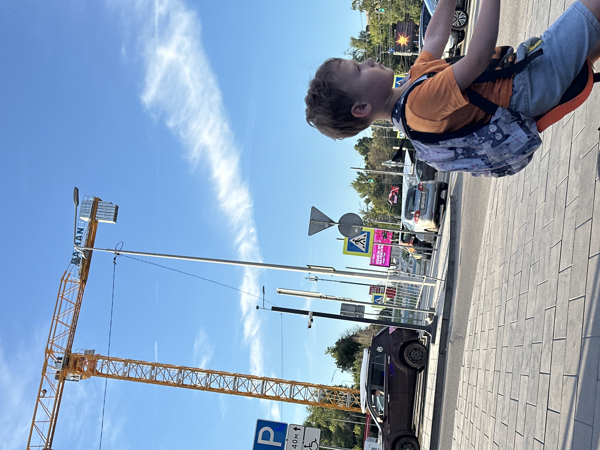 A young boy is pictured looking up towards the sky, with a construction crane dominating the background. He is wearing an orange t-shirt and denim shorts, with a backpack adorned with a playful cat pattern slung over his shoulders. The bright blue sky is dotted with wispy white clouds.
The scene appears to be outdoors, likely in an urban or semi-urban environment, given the presence of traffic lights, road signs, and pavement tiles. A large yellow construction crane is a prominent feature, its towering structure reaching towards the sky. In the foreground, the boy is seated, possibly on a swing or similar apparatus, as suggested by the orange frame visible around him.
A dark-colored car is parked on the right side of the image, and other vehicles are visible in the background, indicating a street or parking area. A sign with Cyrillic text, possibly advertising a store or event, is also visible. The lighting suggests it is daytime, likely sunny. The boy's gaze upwards conveys a sense of curiosity or wonder. The location context provided is Nizhny Novgorod, Russia.