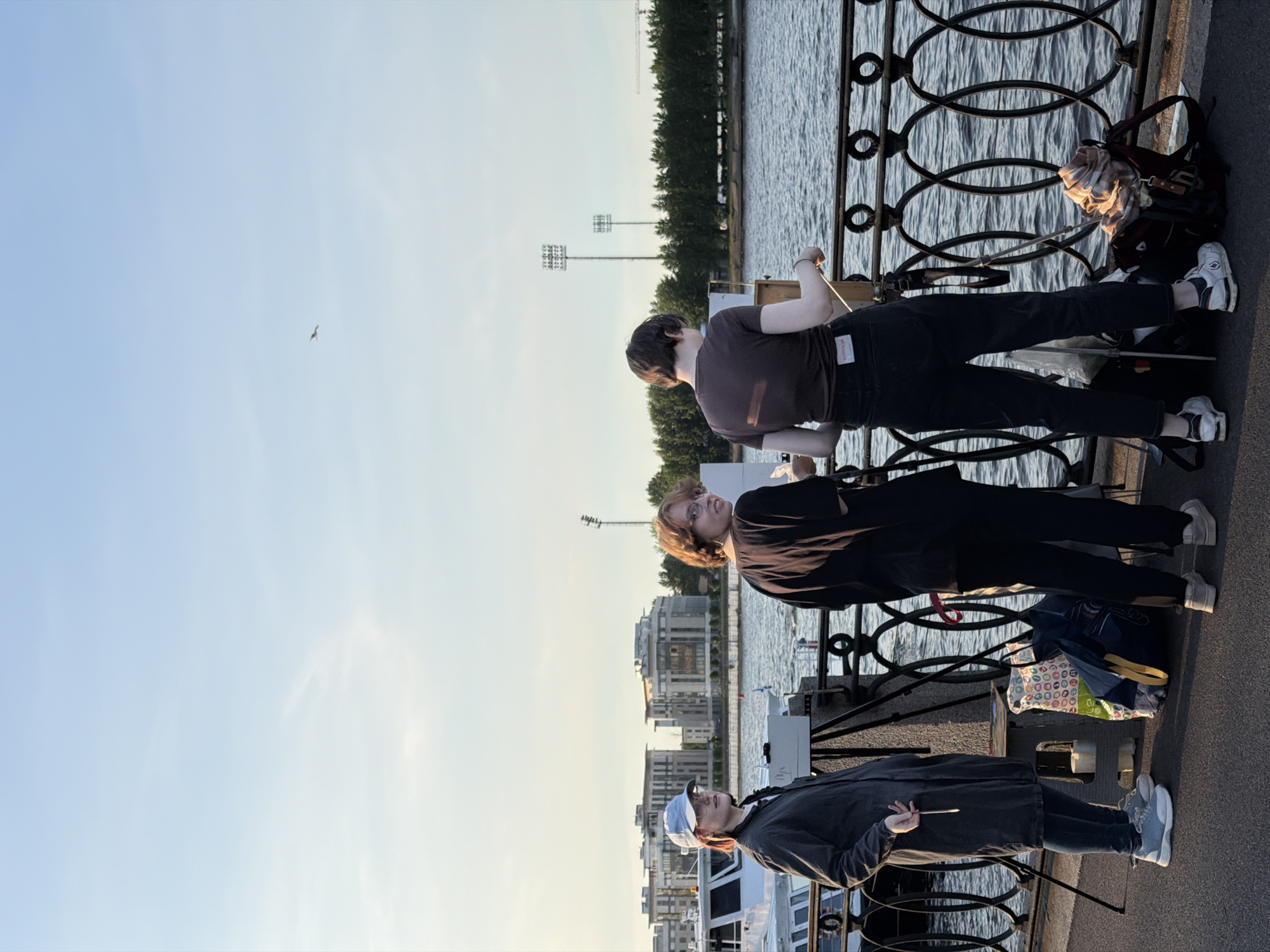 Three young people are standing on a paved walkway next to a decorative metal railing overlooking a body of water. The person on the left, wearing a blue cap, glasses, and a dark gray trench coat, holds a paintbrush. In the center, a person with short, reddish-brown hair and glasses, wearing a black shirt and pants, is looking back over their shoulder. The person on the right, with short dark hair, is wearing a brown t-shirt and dark jeans and is facing away from the camera, holding a paintbrush and looking at an easel. Behind them, across the water, is a cityscape with modern buildings and trees. Two tall light poles are visible in the distance. A boat is docked on the left side of the image. Several bags are on the ground near the people. The sky is a clear, pale blue with a single bird in flight. The scene appears to be outdoors in a public area, likely a promenade or embankment in Saint Petersburg, Russia, given the architectural style and the presence of a large body of water.