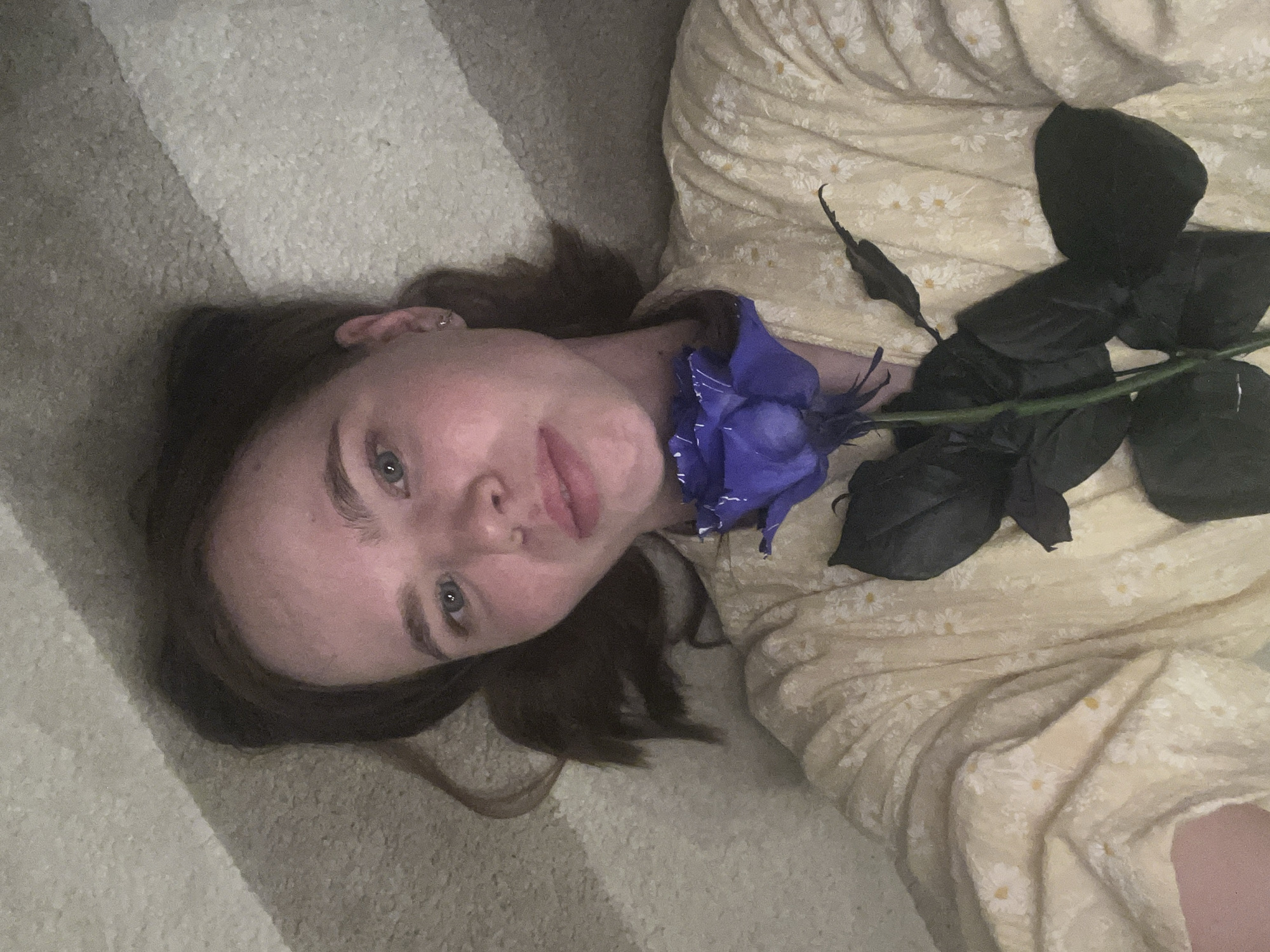 A young Caucasian woman is lying down, looking up at the camera. She has fair skin, light brown hair, and blue eyes. She is wearing a light yellow, floral-patterned dress. A blue rose lies on her chest near her neck. Beside it is a black rose with several dark leaves. The woman is lying on what appears to be a light-colored carpet with horizontal stripes. The setting appears to be indoors. The location and city of Chaikovsky, Russia cannot be confirmed from the image.