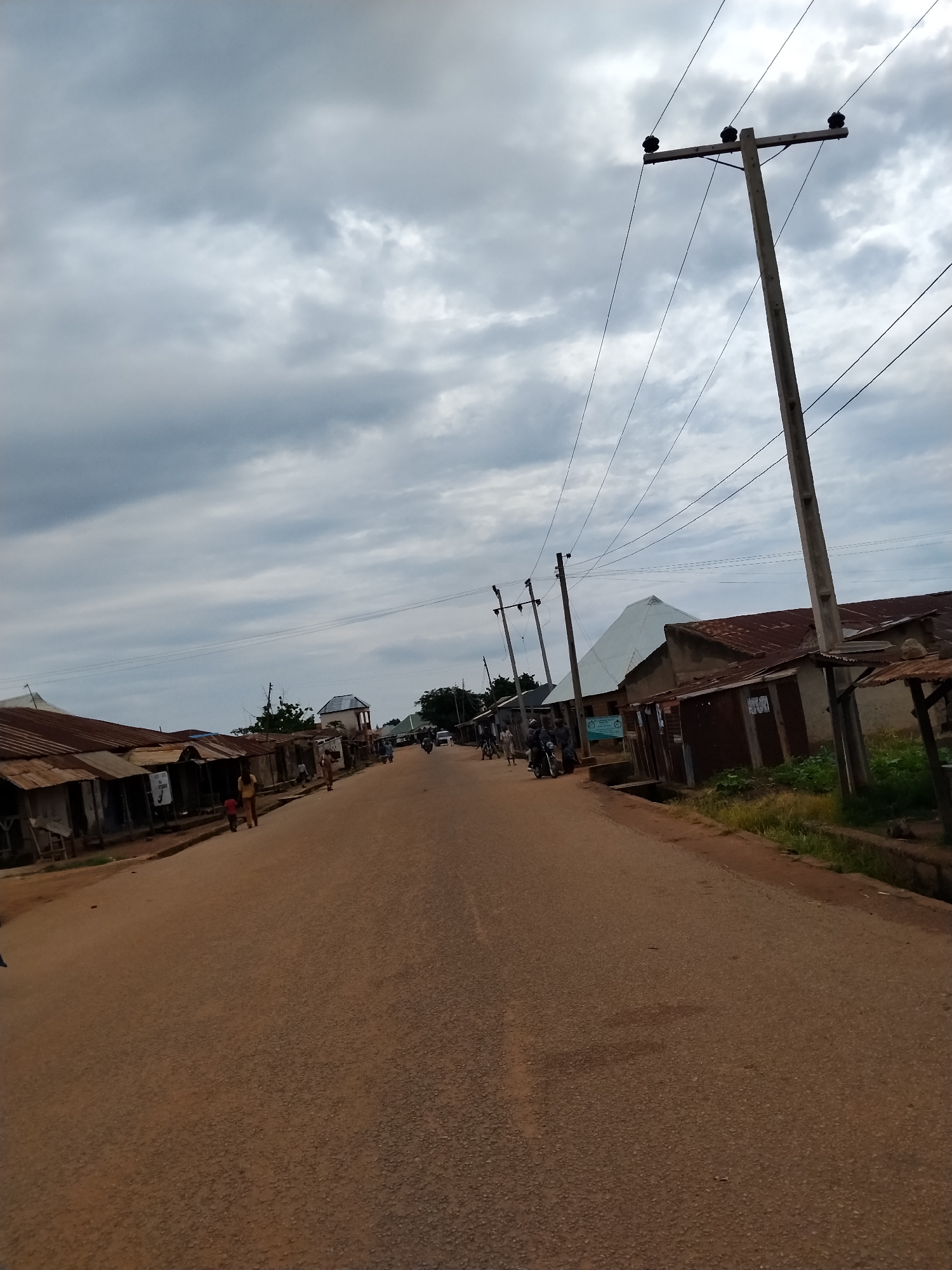 A street scene in Mangu Town, Nigeria, features a dirt road lined with low-rise buildings, many with corrugated metal roofs. Utility poles with power lines run parallel to the road. The sky is overcast with heavy clouds. Several individuals and a motorcycle are visible on the road and sidewalks. The buildings appear to be commercial establishments, some with signs. The overall environment suggests a developing or rural urban setting.