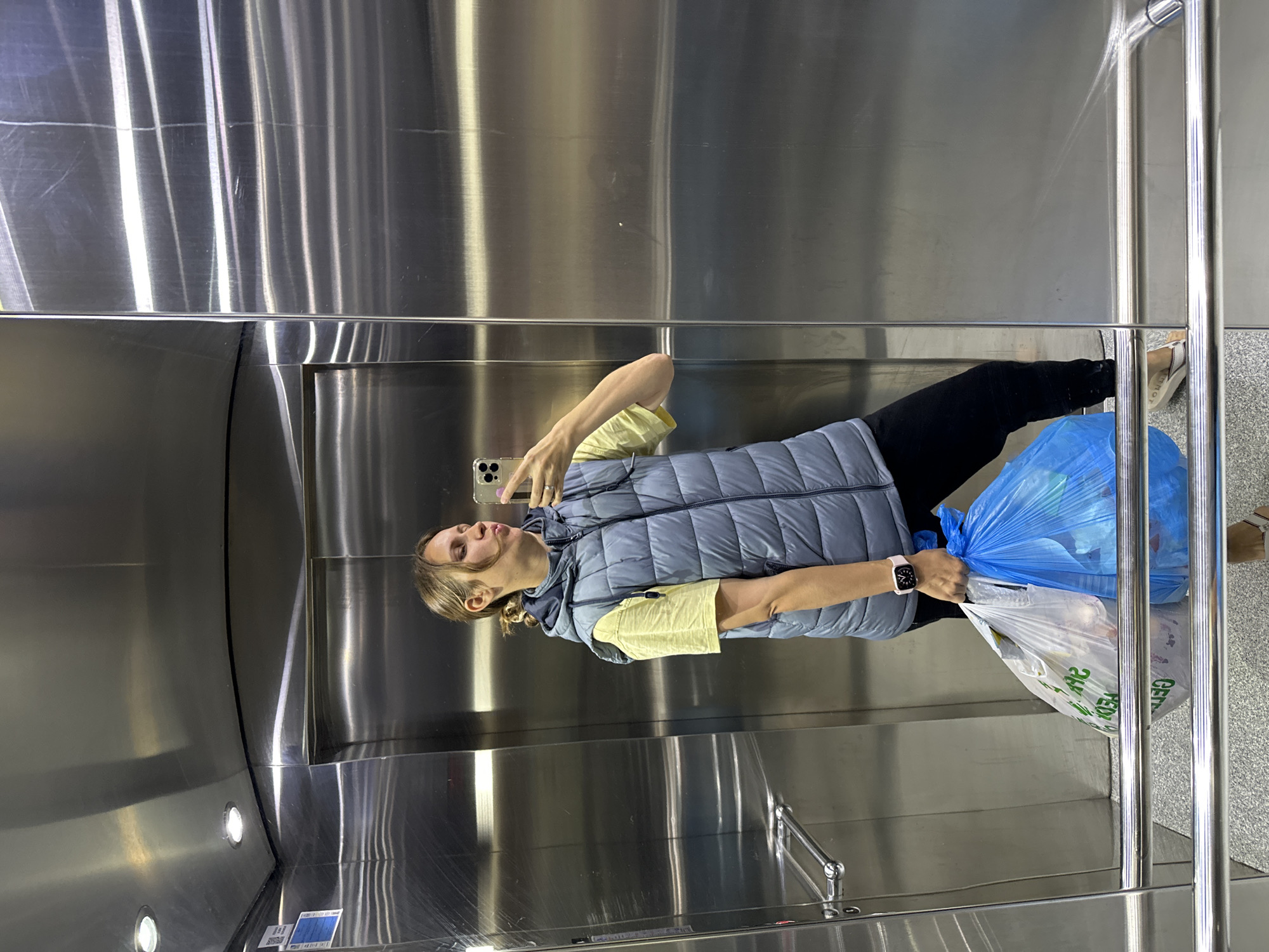 A female is taking a selfie in an elevator. She is holding two bags of trash. The elevator is lined with stainless steel. The location of this image is not available.