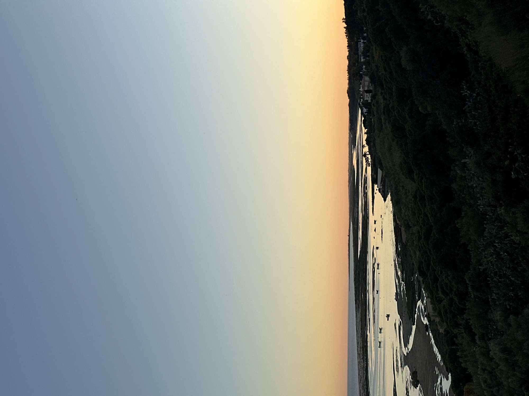 The image shows a landscape at what appears to be dusk or dawn. The sky transitions from a light blue on the left to an orange hue near the center. A body of water runs along the right side of the image, reflecting the light of the sky. The shoreline is bordered by dense vegetation. The location is Vila Real de Santo António, Portugal. Visible are sandy areas and water channels formed by the tide. The scene suggests a coastal environment. No people are visible in the frame.