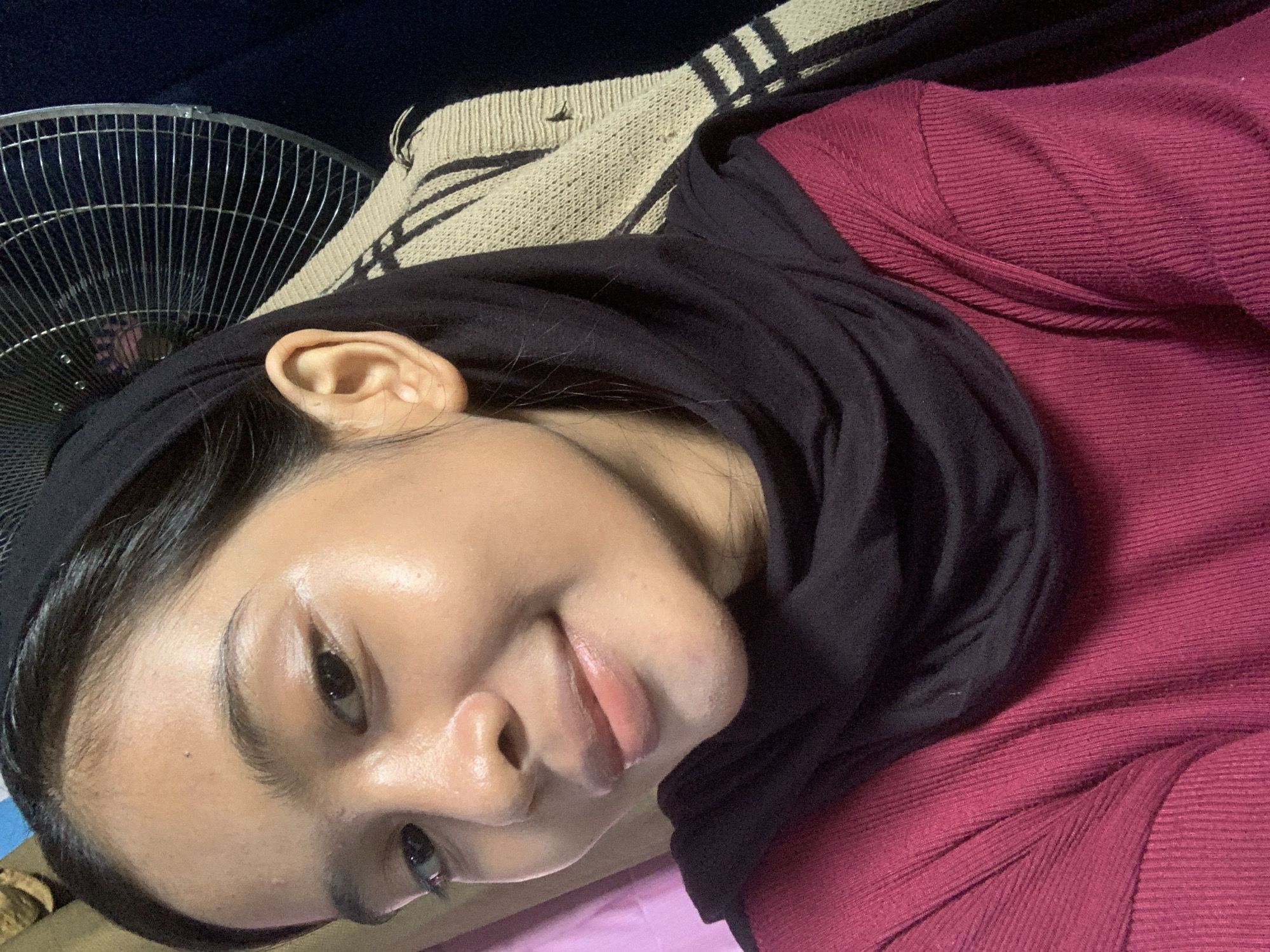 The image is a close-up, selfie-style shot of a young woman with dark hair and brown eyes, wearing a black hijab and a maroon ribbed sweater. Her skin appears to have a dewy, possibly highlighted finish. She is looking towards the camera and smiling gently. In the background, to her right, a metal fan with a wire grill is visible. To her left, a pink, textured surface and a beige and black striped woven blanket or fabric are partially visible. The overall setting appears to be indoors, with a dark background. There is no indication of the location being Sungai Meriam, Indonesia, as the image provides no geographical context.
