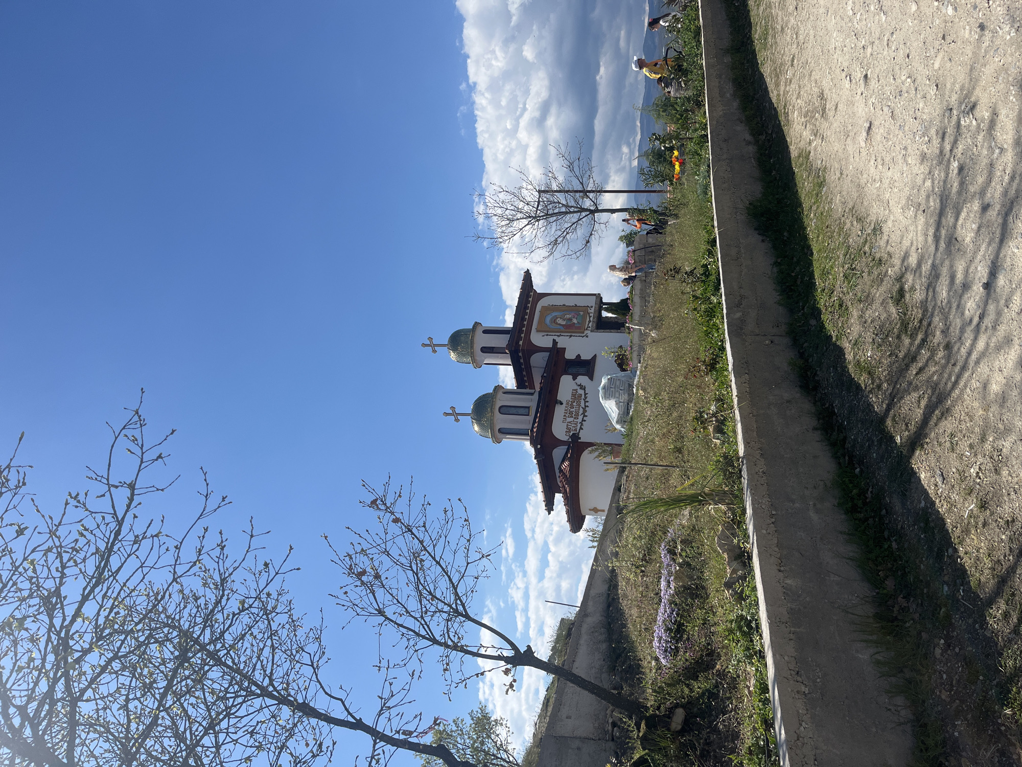 A slightly tilted, medium shot captures a church in Starchevo, Bulgaria against a backdrop of a partly cloudy, bright blue sky. The white church, adorned with twin green onion domes topped with gold crosses, is located behind a short stone wall. Several people stand nearby, some appearing to take pictures. To the side of the frame is a tree without leaves. A dirt path runs along the edge of the shot, partially covered in shadows.