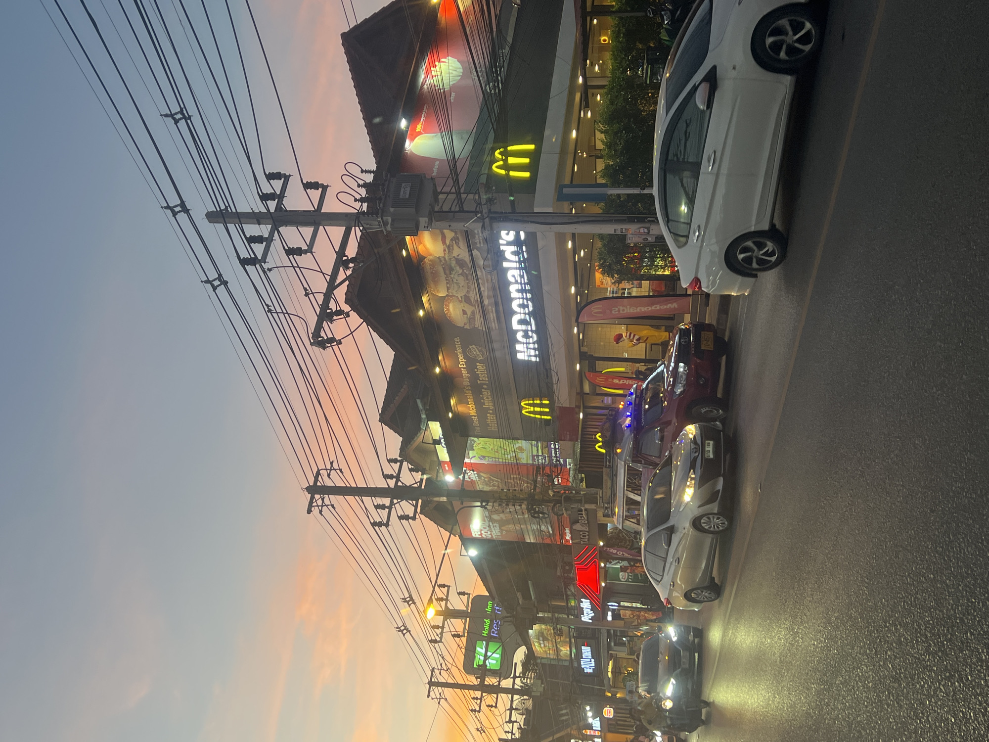 In the city of Ko Samui, Thailand, a street scene unfolds at dusk. A prominent McDonald's restaurant, illuminated by its iconic golden arches and interior lighting, stands on the right side of the image. The restaurant features large advertisements showcasing burgers and menu items. Adjacent to McDonald's are other businesses, including a "Holiday Inn Resort" with a green sign, and others partially visible. The street is lined with numerous utility poles and a complex network of overhead wires. Cars and a motorcycle are present on the asphalt road. In the foreground, a white sedan and a silver sedan are driving, while a red pickup truck is also visible. The sky displays a gradient of soft pastel colors, transitioning from pale blue at the top to shades of pink and orange near the horizon, indicating the sunset.