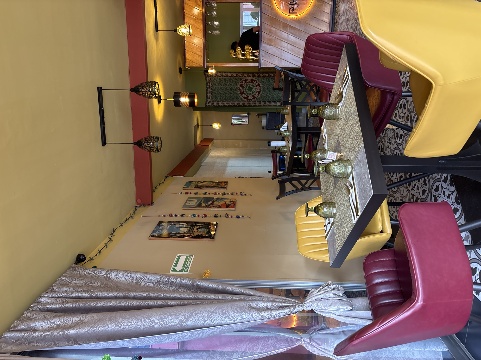 The interior of a restaurant is depicted in the image. A table is centrally located and set with glassware and cutlery. Yellow and red chairs surround the table. A window is draped with a patterned curtain. The walls are a muted yellow color with a red accent strip near the ceiling. String lights and framed artwork adorn the walls. Lighting fixtures are suspended from the ceiling. The restaurant appears to be in San Andrés Cholula, Mexico, based on the wall text "Ruta de Evacuación", in Spanish. In the background, another person can be seen in the kitchen area.