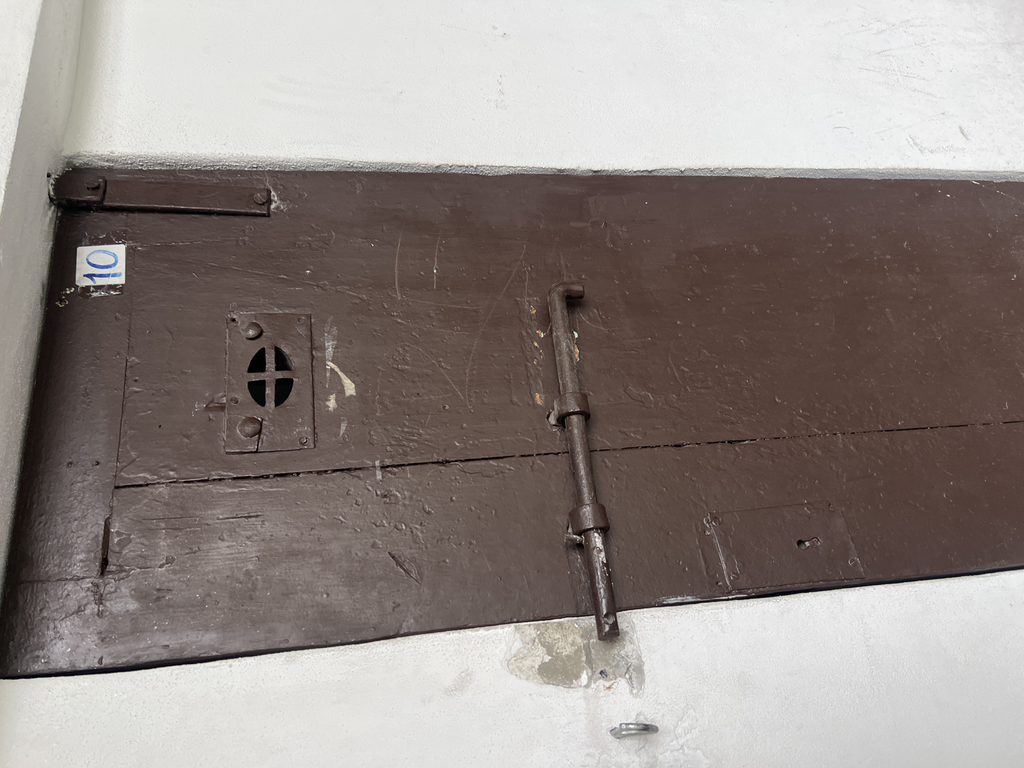The image shows a close-up of a wooden door painted in brown, likely an access hatch or similar. The door is weathered and shows signs of age, with visible cracks and scratches on the painted surface. A metal slide bolt mechanism secures the door, and a small square viewing port with a cross-shaped opening is visible near the upper left corner of the door. A number "10" is painted in blue on the upper left corner of the door. The door is set into a white wall, with some areas of discoloration and patch work on the wall surface near the bottom edge of the door. The image was taken in the city of Puebla, Mexico.