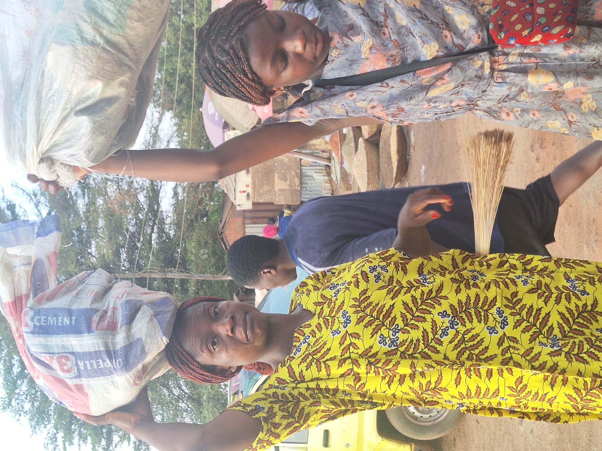 The image shows two women in Kakuri Gwari, Nigeria. One woman is wearing a yellow dress with a leaf pattern, carrying a bag of cement on her head and holding a broom. The bag reads "CEMENT OPELLA 3x". Another woman, wearing a floral dress, is holding a white sack overhead. There are other people nearby, including a person in a blue shirt. The background includes trees, buildings, and a vehicle.