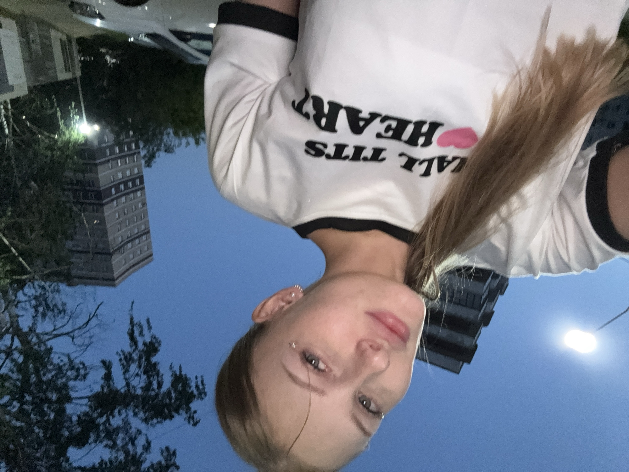 In a selfie taken outdoors at dusk, a young woman with blonde hair and facial piercings poses in front of two buildings. She is wearing a white t-shirt with black trim that has the words "ALL TITS HEART" printed in black lettering, with a pink heart symbol between "ALL" and "TITS". The background includes a street lamp, trees, and a white car parked on the street. The image is set in a city, likely in Perm, Russia, given the architectural style of the buildings.
