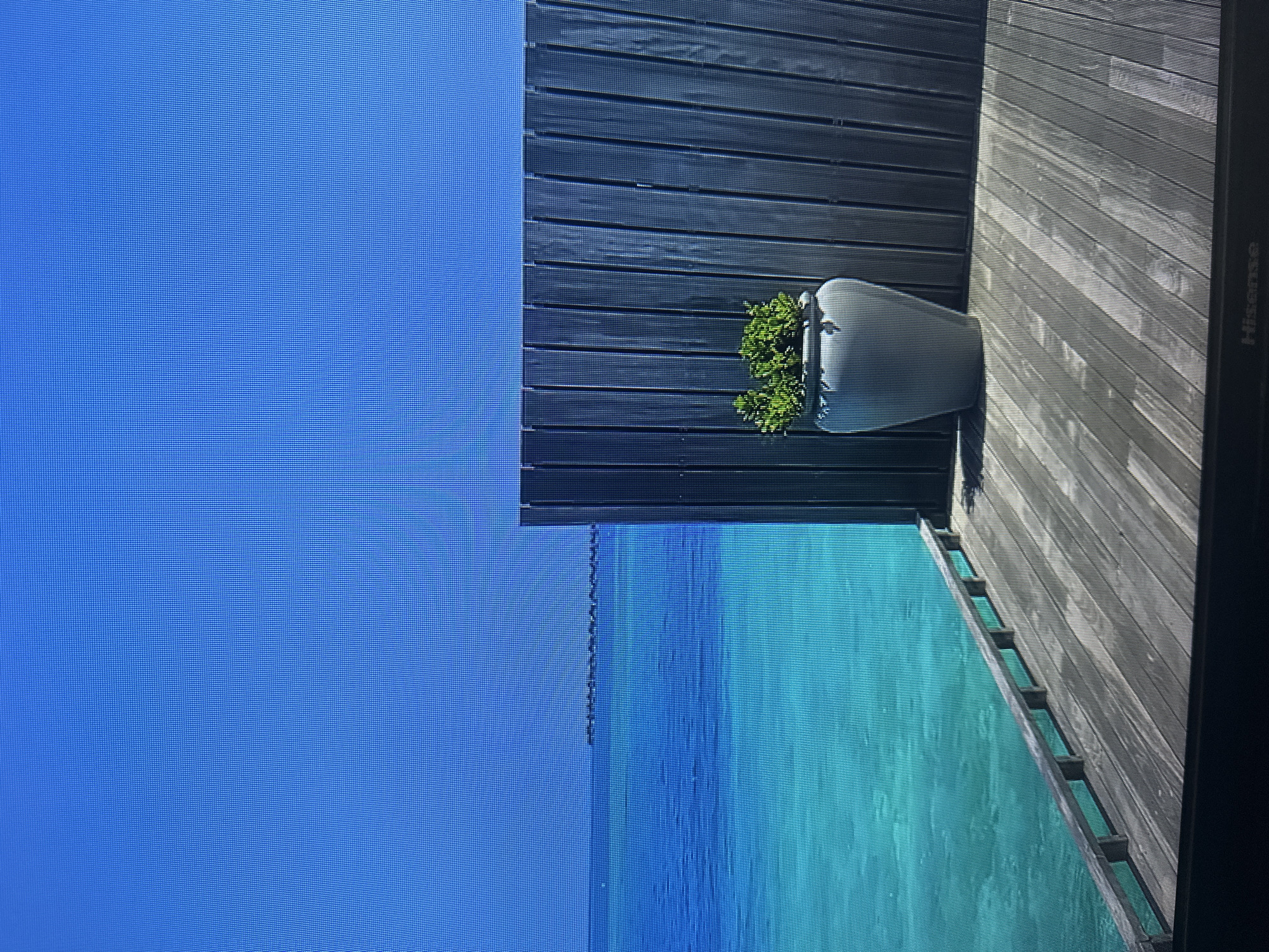 The image depicts a serene scene with a bright blue sky and clear turquoise water. A wooden deck extends into the water, and a dark wooden fence runs along the right side of the frame. A large, light gray planter sits on the deck, containing a vibrant green shrub. In the distance, a row of small structures or buildings can be seen on the horizon. The scene suggests a tropical or coastal location, likely a resort or private villa. The image does not contain any information that would indicate it is located in Kyiv, Ukraine.