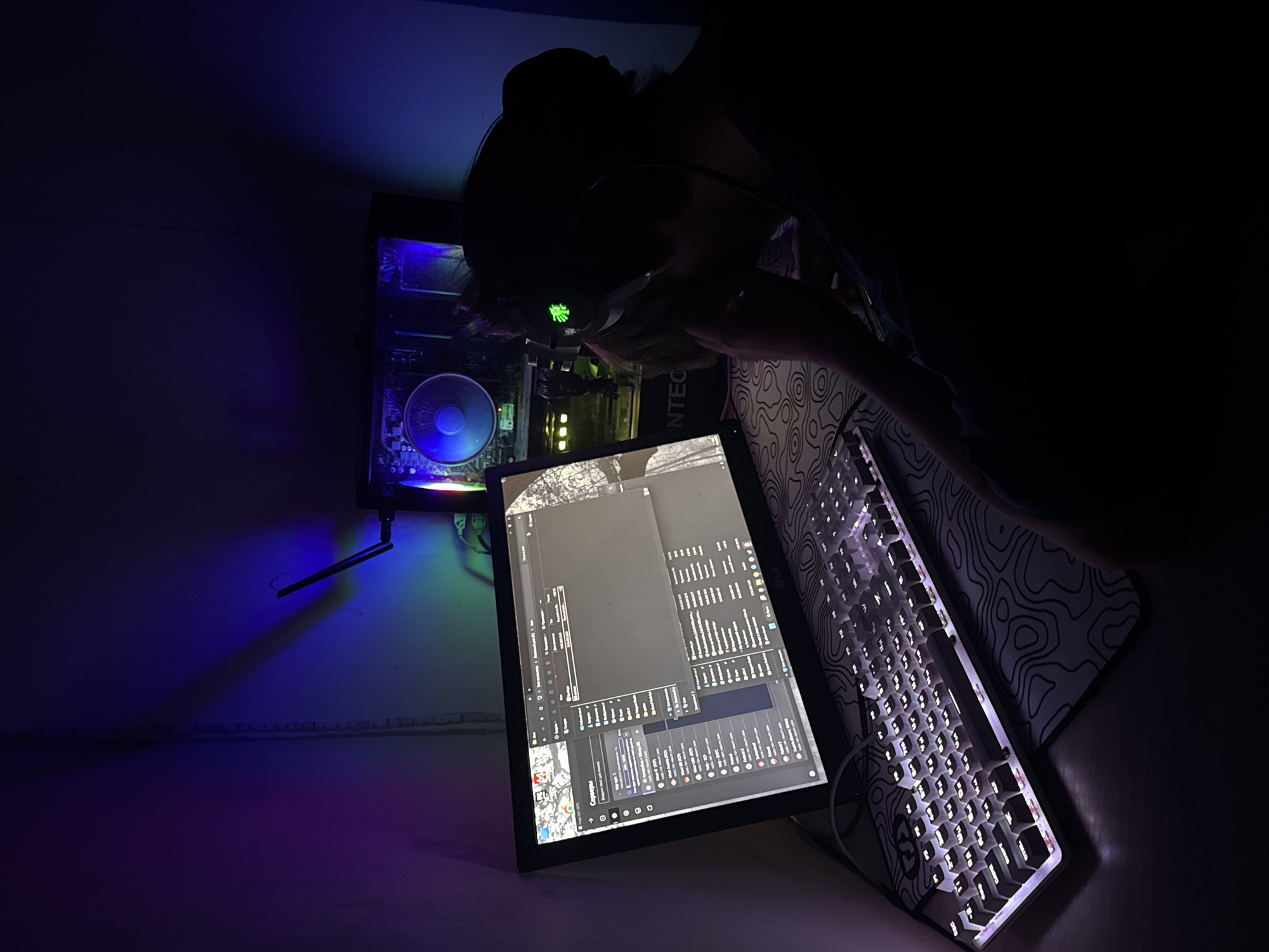 In a dimly lit room, likely a bedroom or home office in Khomutovo, Russia, a person wearing headphones is seated in front of a computer. The scene is characterized by the glow of computer monitors and ambient lighting, casting purple and blue hues on the walls. The computer setup includes a large monitor displaying what appears to be a file explorer or server list, a keyboard with illuminated keys, and a visible computer tower with a fan. The person's posture suggests focused engagement with the computer, possibly gaming or working. The overall environment is indicative of a personal space designed for digital interaction.