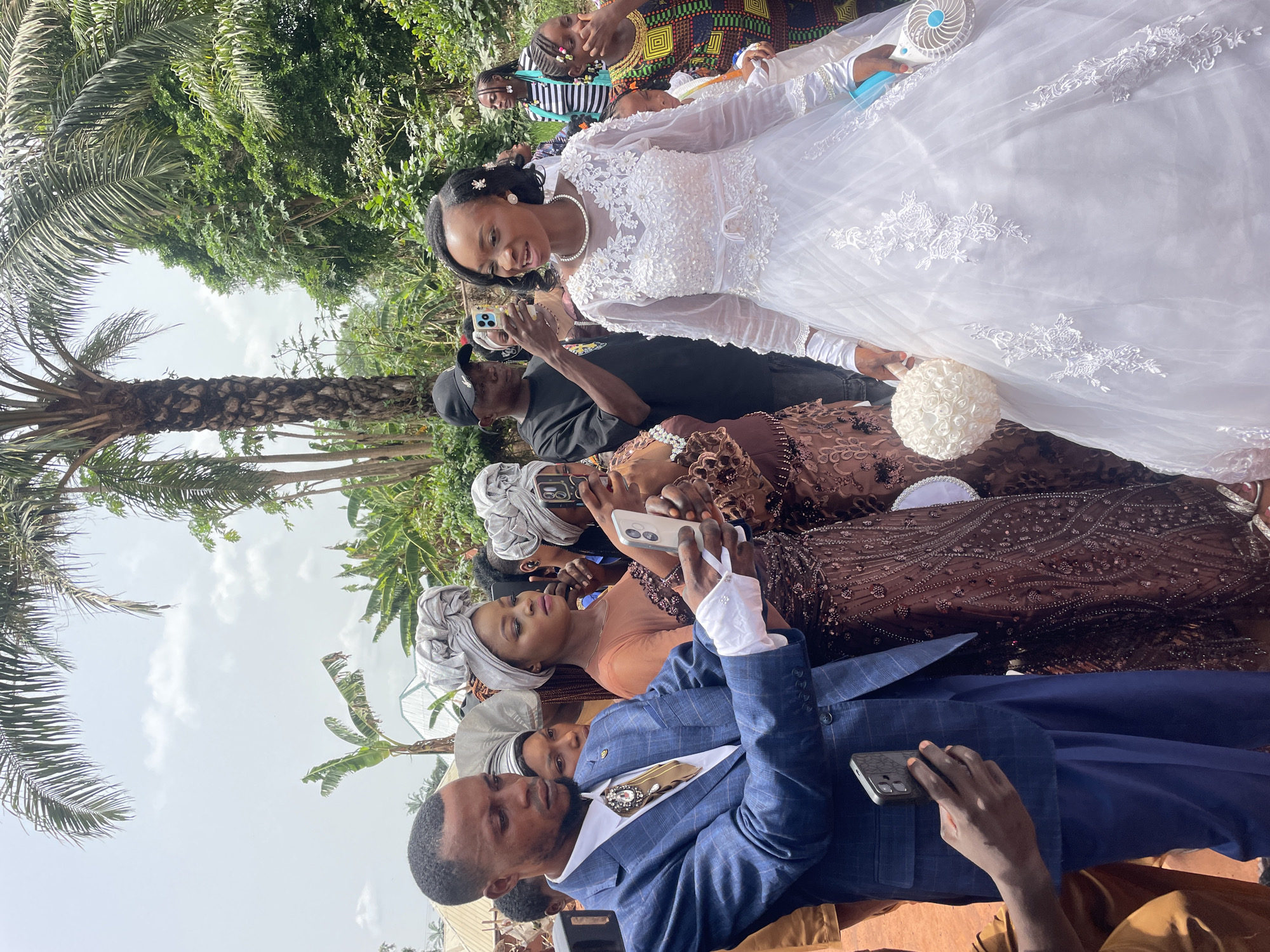 A medium shot depicts a wedding scene in Ogbe, Nigeria. The bride is dressed in a white gown with lace detailing and a pearl necklace. She is surrounded by people who are taking photos of her with their cellphones. A man in a blue suit is in the foreground, also holding a phone. The background shows palm trees and other green foliage, with a cloudy sky visible.