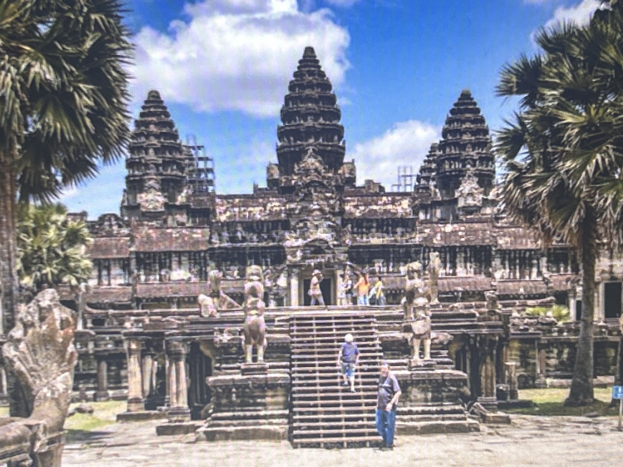The image depicts a large, ancient stone temple complex, likely Angkor Wat in Cambodia, under a bright blue sky with scattered clouds. The temple features multiple tiered towers and elaborate carvings. In the foreground, a wide set of stone stairs leads up to the main structure. Several people are visible: a man in a blue shirt and jeans stands at the bottom right of the stairs, looking towards the temple; another person in a blue shirt and shorts is ascending the stairs; a group of people are gathered on a platform near the entrance of the temple; and two figures, possibly statues, stand on either side of the base of the stairs. Palm trees frame the left and right sides of the temple. The ground in front of the temple appears to be paved or compacted earth. The scene is brightly lit, suggesting daytime. The location is not in Sofia, Bulgaria.