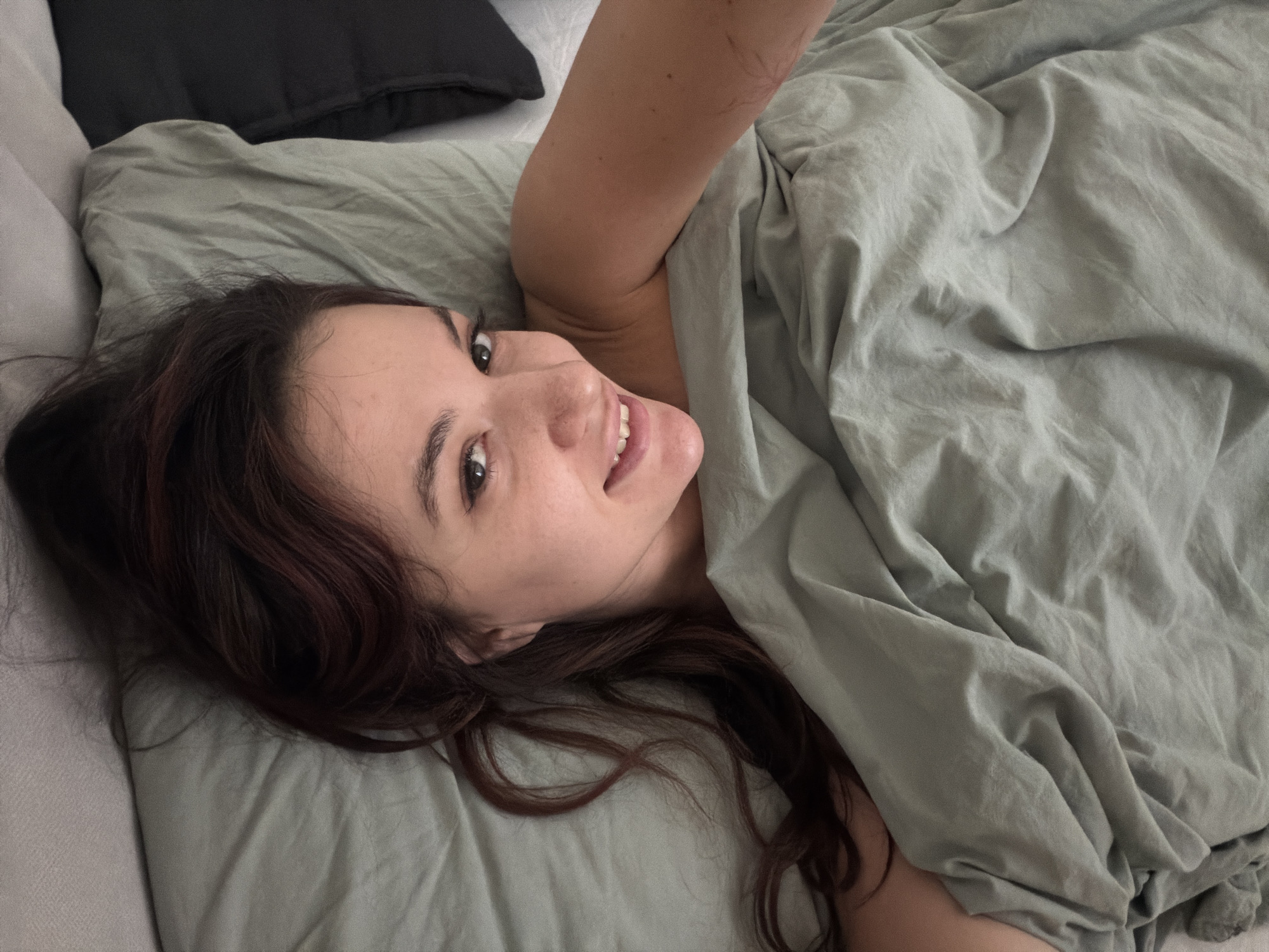 A young woman is lying in bed, partially covered by a light green duvet. Her head is on a matching light green pillow. She has long, wavy dark brown hair and is smiling. A black pillow is visible near the top left corner of the frame. Part of an arm is visible above the duvet. The location is not identifiable as Belgrade, Serbia.
