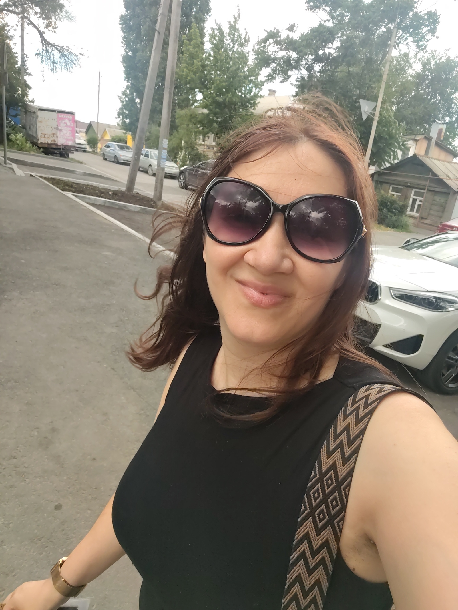 The image is a selfie taken by a woman with brown hair, wearing large, dark sunglasses and a black sleeveless top. She has on a gold-colored bracelet on her left wrist and a bag with a patterned strap slung over her right shoulder. The woman is smiling directly at the camera, and her expression suggests a cheerful mood.
The scene behind her depicts a street in what appears to be a residential area. Several cars are visible, including a white SUV in the foreground and other vehicles parked or driving in the background. There is a rustic-looking wooden house with a window, and further back, more houses and trees can be seen. Two utility poles stand prominently in the middle ground. The overall setting suggests an outdoor environment, possibly during the daytime, as indicated by the lighting. A truck with some writing on its side is parked on the left side of the image.
The weather appears to be overcast, with a bright, diffused light. The woman's hair is slightly windswept, suggesting a gentle breeze. The presence of trees and houses indicates an urban or suburban setting. The writing on the truck is partially visible, but not legible enough to decipher specific details. The overall impression is of a casual, everyday moment captured outdoors.