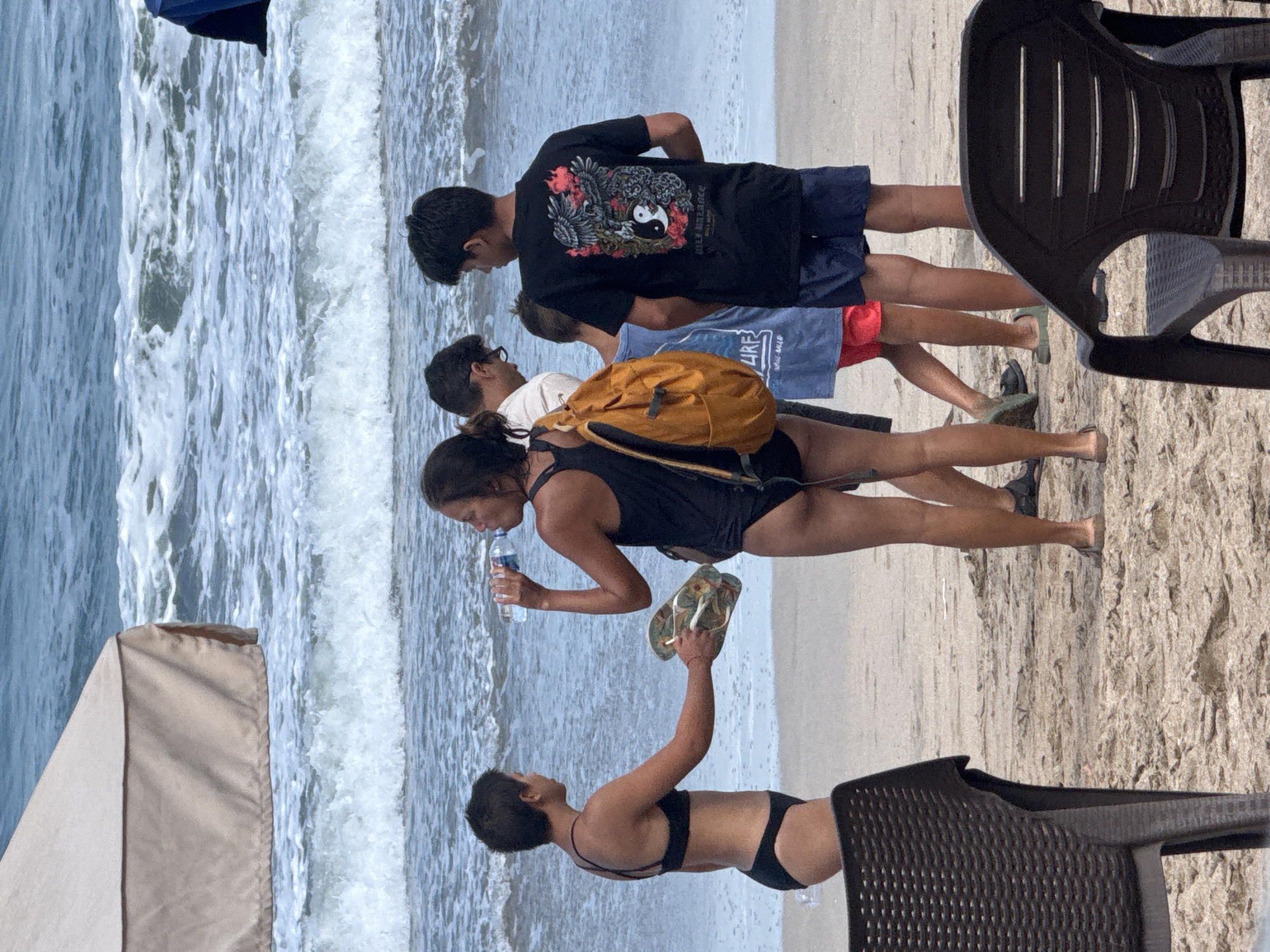 In Kuta, Indonesia, a group of people are gathered on a sandy beach near the ocean. The water is a vibrant blue with white-capped waves, suggesting a sunny day with active surf. The beach itself is sandy, with some scattered rocks and what appears to be a black plastic chair in the foreground, hinting at a recreational area.
The subjects in the image include several individuals. Closest to the camera, a person wearing a black bikini is holding a pair of flip-flops in one hand and reaching out with the other. Behind them, a woman in a black swimsuit is holding a bottle of water to her lips, with an orange backpack slung over her shoulder. Further back, several individuals in casual beachwear, including shorts and t-shirts, are standing. One person wears a black t-shirt with a graphic design featuring an eagle and a yin-yang symbol. The people appear relaxed, likely enjoying a day at the beach. The presence of a beach umbrella to the left suggests the area is set up for sunbathers. The image captures a moment of leisure and outdoor activity on a tropical beach.