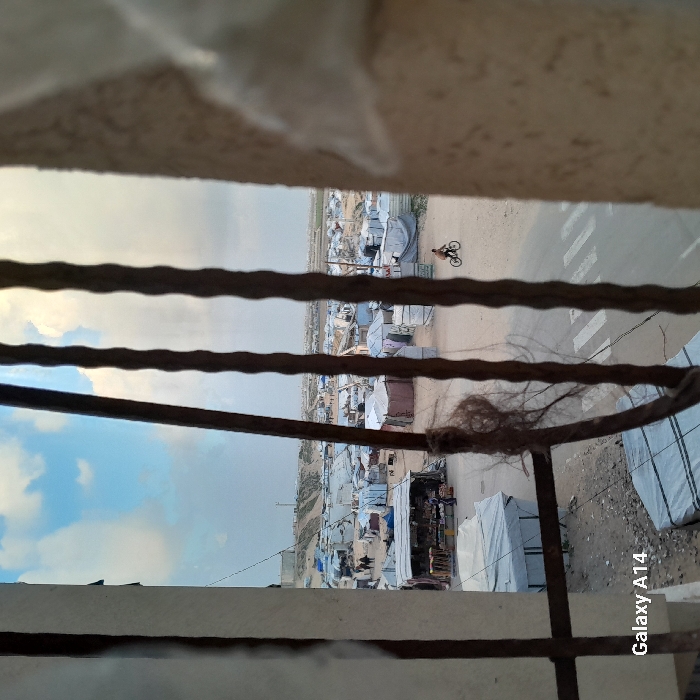 The image, displayed rotated 90 degrees clockwise, captures an outdoor scene from an elevated vantage point, looking down through a foreground barrier. If viewed in its natural orientation (rotated 90 degrees counter-clockwise), the scene depicts a wide panorama of an informal settlement or camp under a partially cloudy sky.
In the immediate foreground, blurring the top and bottom of the frame and extending horizontally across the middle, are several thick, dark, weathered bars or ropes. These appear to be part of a railing, fence, or window security bars. The topmost and bottommost blurred sections suggest a concrete or stucco wall or frame. On the rightmost of these horizontal bars, a tangled mass of light brown, fibrous material, resembling matted hair or natural fibers, is visible.
Beyond these foreground elements, stretching into the mid-ground and background, lies a densely packed settlement. It consists of numerous makeshift structures, tents, and small, simple buildings constructed from various materials such as corrugated metal, tarpaulins (some blue, some white), and possibly other scrap materials. The structures appear to be closely clustered together, indicating a crowded living environment.
A dirt road, or a well-worn unpaved path, weaves through the settlement. On this road, white parallel lines are painted, forming what appears to be a pedestrian crosswalk. A single person is seen riding a bicycle along this road, moving from left to right in the mid-ground. Near the road, some structures appear to have small market stalls or areas where goods might be displayed.
In the far background, beyond the settlement, the terrain appears relatively flat and barren, possibly a dusty plain or arid landscape. The sky, visible above this distant horizon, is partly cloudy, with patches of blue sky showing through light grey clouds. The overall lighting suggests it could be late afternoon or an overcast day, as there are no harsh shadows.
The only discernible text in the image is a watermark, "Galaxy A14," positioned vertically along the far right edge of the image as it is currently displayed.
