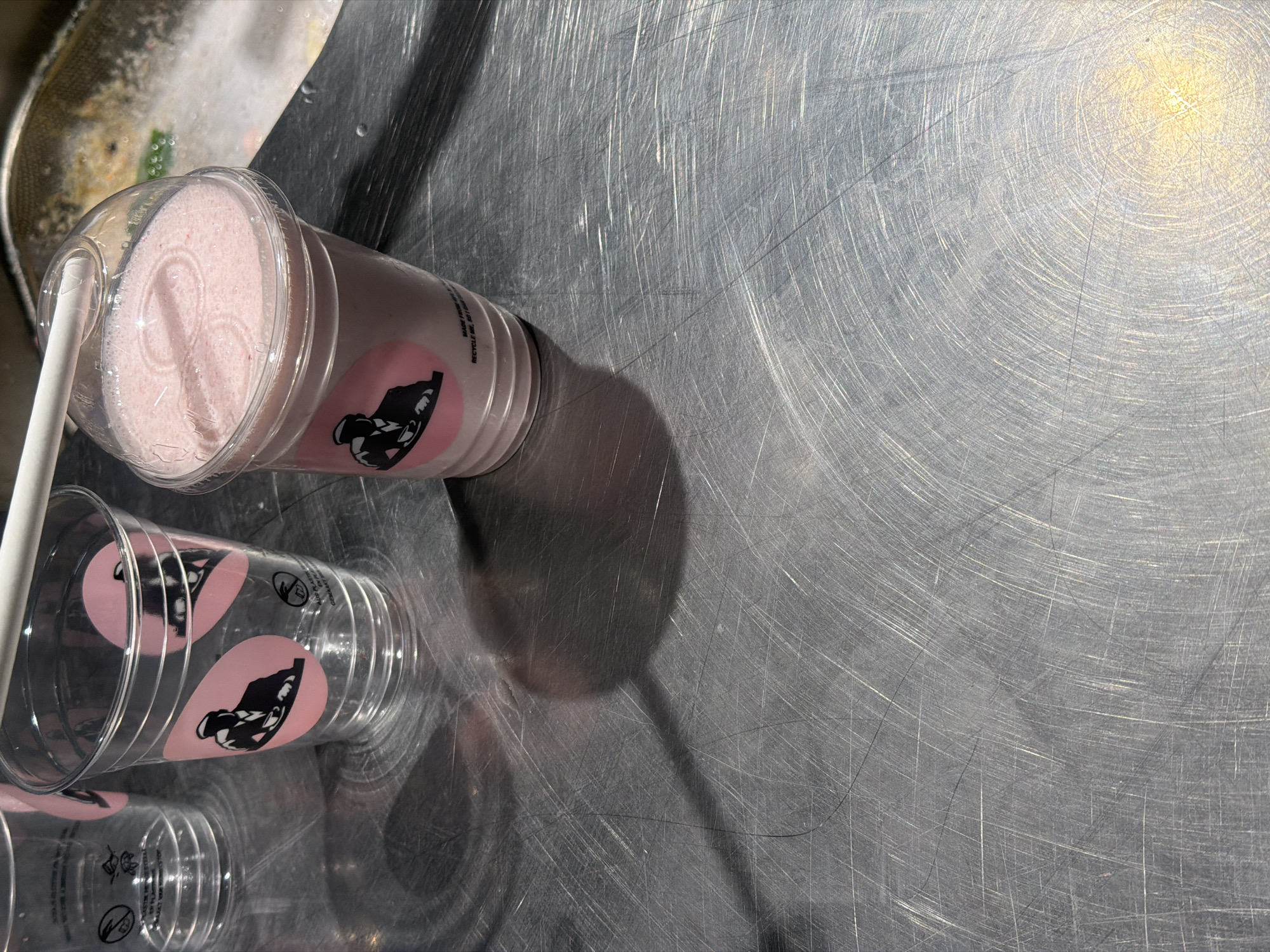 The image depicts a stainless steel surface on which a full cup of a pink beverage with a white straw stands. There are two empty cups stacked next to it, all with the same logo. The surface shows a pattern of concentric scratches and scuffs, suggesting a well-used industrial or commercial surface, possibly a kitchen or food preparation area. Given the logo on the cups, this could be from a location of a business associated with that logo, potentially in Paris, France. The lighting suggests artificial illumination with a bright, direct light source. There is no indication of people or ongoing interactions in the immediate view.