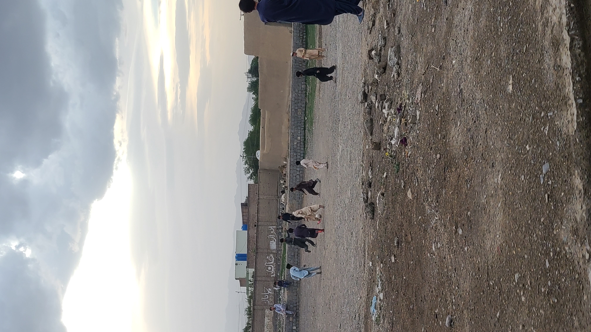 The image depicts a group of men and boys playing soccer (football) on a dusty, unpaved field in Qilla Saifullah, Pakistan. The scene appears to be set in the late afternoon or early evening, indicated by the bright, low-angled sunlight breaking through the clouds, creating a strong glare in the sky.
In the foreground and mid-ground, several individuals, mostly wearing traditional shalwar kameez attire, are engaged in the game. One person is positioned near a soccer ball. The ground is rough and stony, with some scattered debris and litter visible on the right side.
In the background, a row of low-rise buildings, appearing to be constructed from mud-brick or similar earthy materials, line the edge of the field. Beyond these structures, distant mountains are visible under a cloudy sky. The sky transitions from darker, more solid clouds on the left to bright, sunlit areas on the right.
On one of the walls in the mid-ground, some text is visible, possibly in Urdu or Pashto. Parts of words like "خانہ" (khana, meaning house or place) and "ارزاق" (arzaq, meaning sustenance or livelihood) can be discerned, along with other less clear inscriptions. The overall atmosphere suggests a casual, community gathering for recreation in a rural or semi-urban setting.