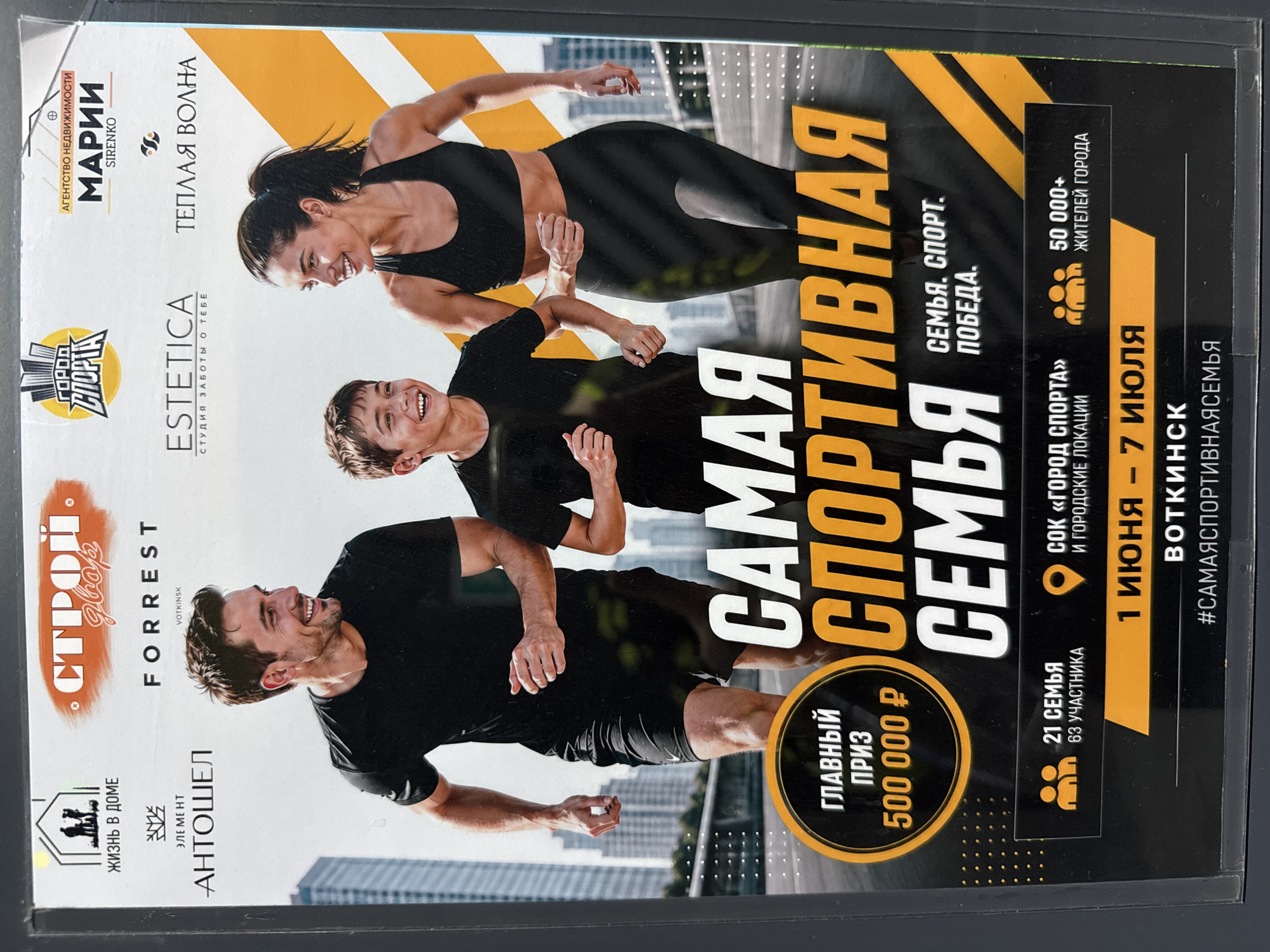 The image is a flyer advertising a sports competition called "The Most Athletic Family" in Votkinsk, Russia, from June 1st to July 7th. The main prize is 500,000 rubles. The competition is sponsored by several local businesses, including a real estate agency (Marii Sirenko), a beauty studio (Estetica), a construction supplies store (Stroy Dvor), and a home goods store (Antoshel).
The flyer features a photo of a smiling family – a man, woman, and child – jogging. The location is referred to as "SOK 'City of Sports' and city locations." There were 21 families with 63 participants in total. The town has over 50,000 citizens. The flyer’s hashtag is #samayasportivnayasemya.