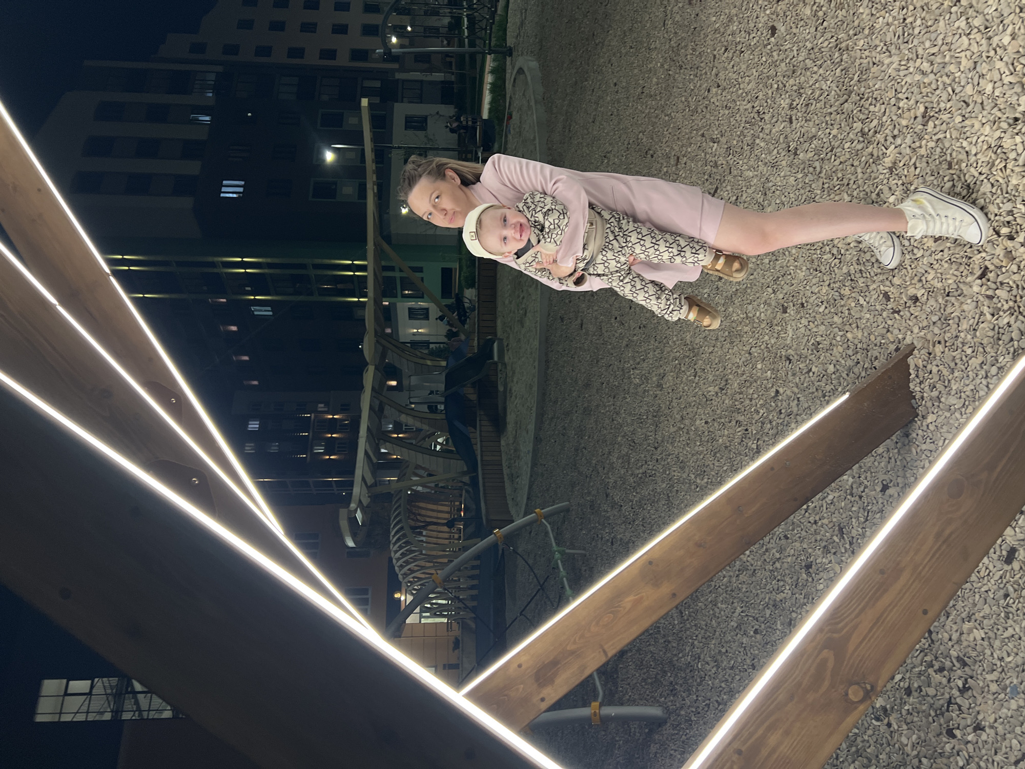 A woman in a pink dress and white sneakers holds a child in a patterned outfit and a white hat in an outdoor setting at night. The location appears to be a playground or public space in a city, possibly Pervomaysky, Russia, based on the prompt. The background features a tall building with lighted windows and playground equipment. The foreground shows a modern wooden structure with embedded LED lights. The ground is covered in small gray stones. The woman is standing, and the child is in her arms. They are looking towards the camera.