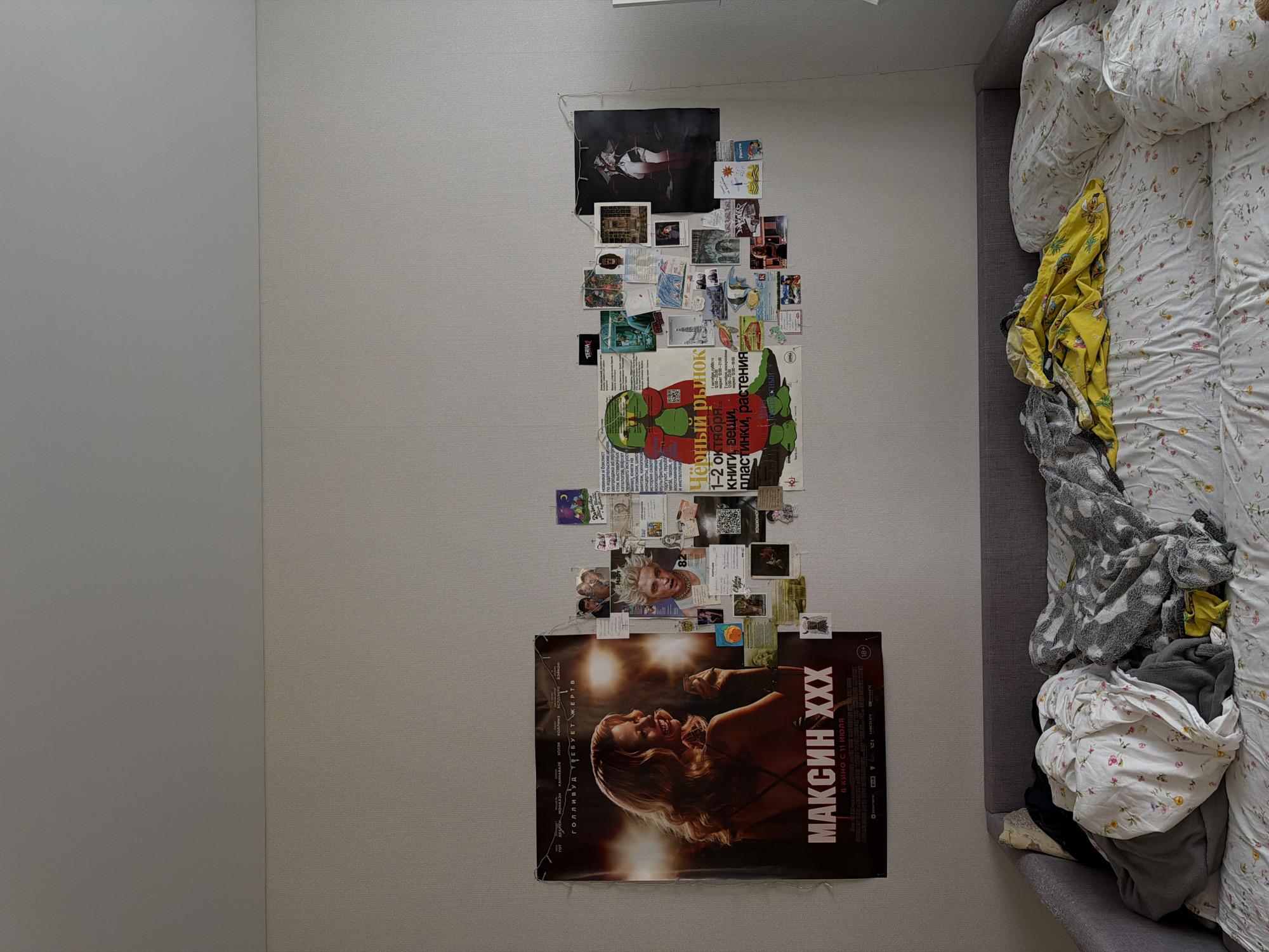 The image presents an overhead view of a room, likely a bedroom, featuring a decorated wall and an adjacent unmade bed. The wall, covered in light grey textured wallpaper, is adorned with a large collage of posters, photos, and printouts.
At the bottom of the wall, there is a prominent movie poster for "МАКСИН XXX" (Maxine XXX), depicting a laughing woman with light hair against a dark, warm-toned background. The poster indicates "В КИНО С 11 ИЮЛЯ" (IN CINEMA FROM JULY 11) and carries an "(18+)" age restriction. Smaller credits like "ГОТ ДЕВИКИ МОНАКЭН КАННАВАЛЕ ХОЛЗИ КОЛЛИНЗ" and phrases such as "ГОЛЛИВУД ТРЕБУЕТ ЖЕРТВ" (HOLLYWOOD DEMANDS VICTIMS) are also visible.
Above the movie poster, a dense collage of various smaller images, postcards, and textual printouts forms an irregular vertical strip. In the middle section of this collage, a large green and red poster advertises "Чёрный рынок" (Black Market) for "1-2 октября" (October 1-2) from "12:00-21:00". It lists items such as "книги, вещи, пластинки, растения" (books, things, records, plants) and includes "ТПЕНЗА", possibly referring to the city Penza. Near the top of the wall display, a dark poster shows a figure in a white outfit.
To the right of the wall, an unmade bed with a grey upholstered frame is visible. The bed is covered with a white duvet featuring a subtle floral pattern. Several pillows are present, and various crumpled textiles, including a yellow patterned blanket and a grey patterned blanket, are tossed on the bed.
The scene suggests a personal, lived-in space. The text in Russian indicates a location in Russia, specifically Засечное as provided by the context. No specific time of day or outdoor weather is discernible.