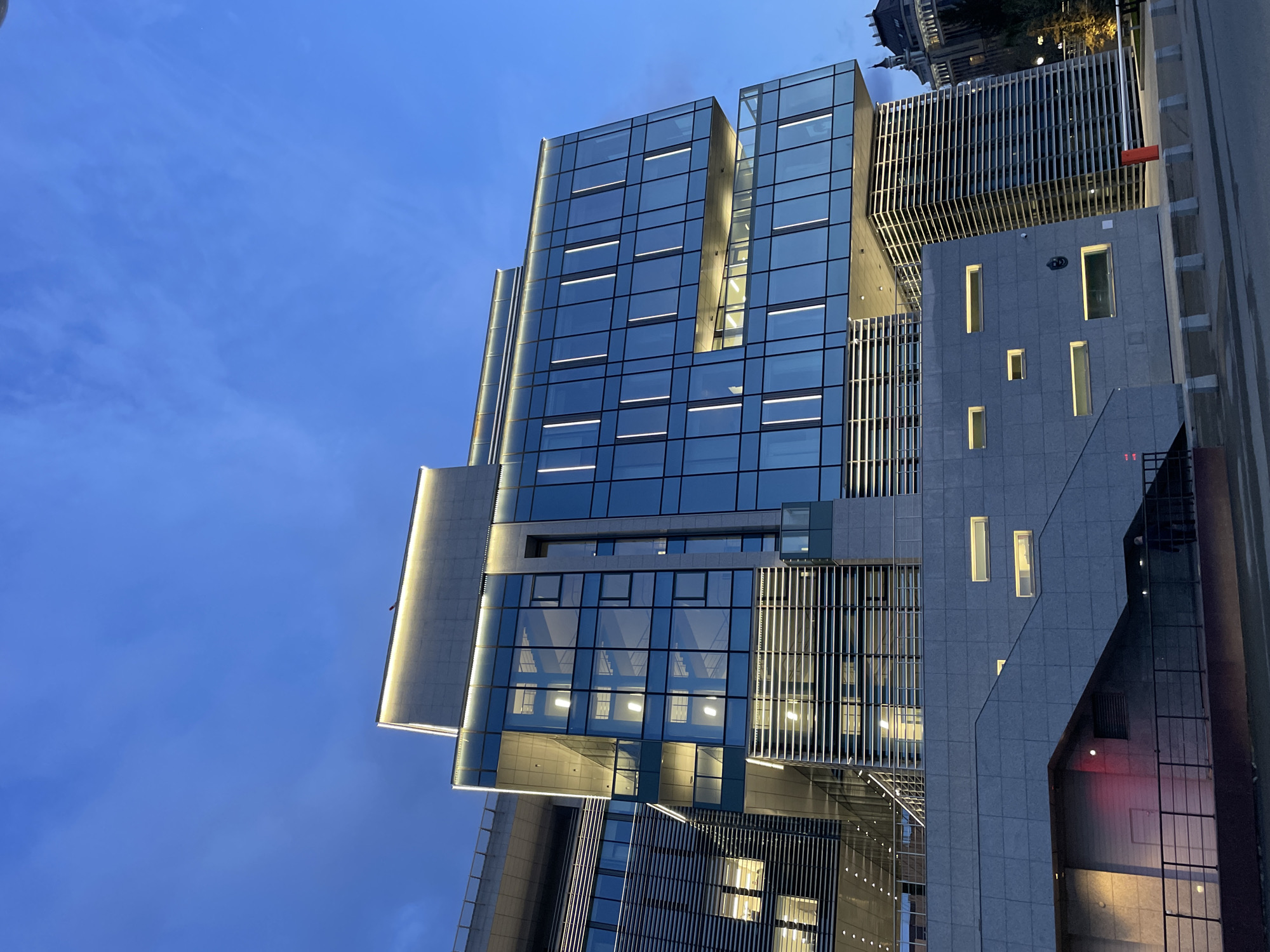 The image displays a striking modern architectural building captured during late twilight or early evening. The main structure is a multi-story edifice characterized by a prominent glass facade that reflects the deep blue sky. Horizontal strip lights are visible within many of the glass panels, illuminating the interior spaces. Adjacent to this glass section, a contrasting part of the building features grey stone cladding with an irregular pattern of illuminated rectangular windows.
The building's design showcases clean lines and a contemporary aesthetic, incorporating both smooth glass and textured stone materials. Several sections feature metallic vertical fins or louvers, adding depth to the facade. Accent lighting along ledges and architectural details highlights the building's contours against the dimming sky. The sky is a clear, vibrant blue with faint, wispy clouds, indicating fair weather. In the upper background, a glimpse of an older building with a dark, possibly domed roof can be seen, suggesting an urban setting where historical and modern architecture converge.
No people or discernible activities are visible in the scene. The illuminated windows and interior lights suggest the building is operational or prepared for evening use. There is no visible text or signage to provide specific location cues. The overall impression is one of sophisticated urban architecture beautifully lit as day transitions into night.