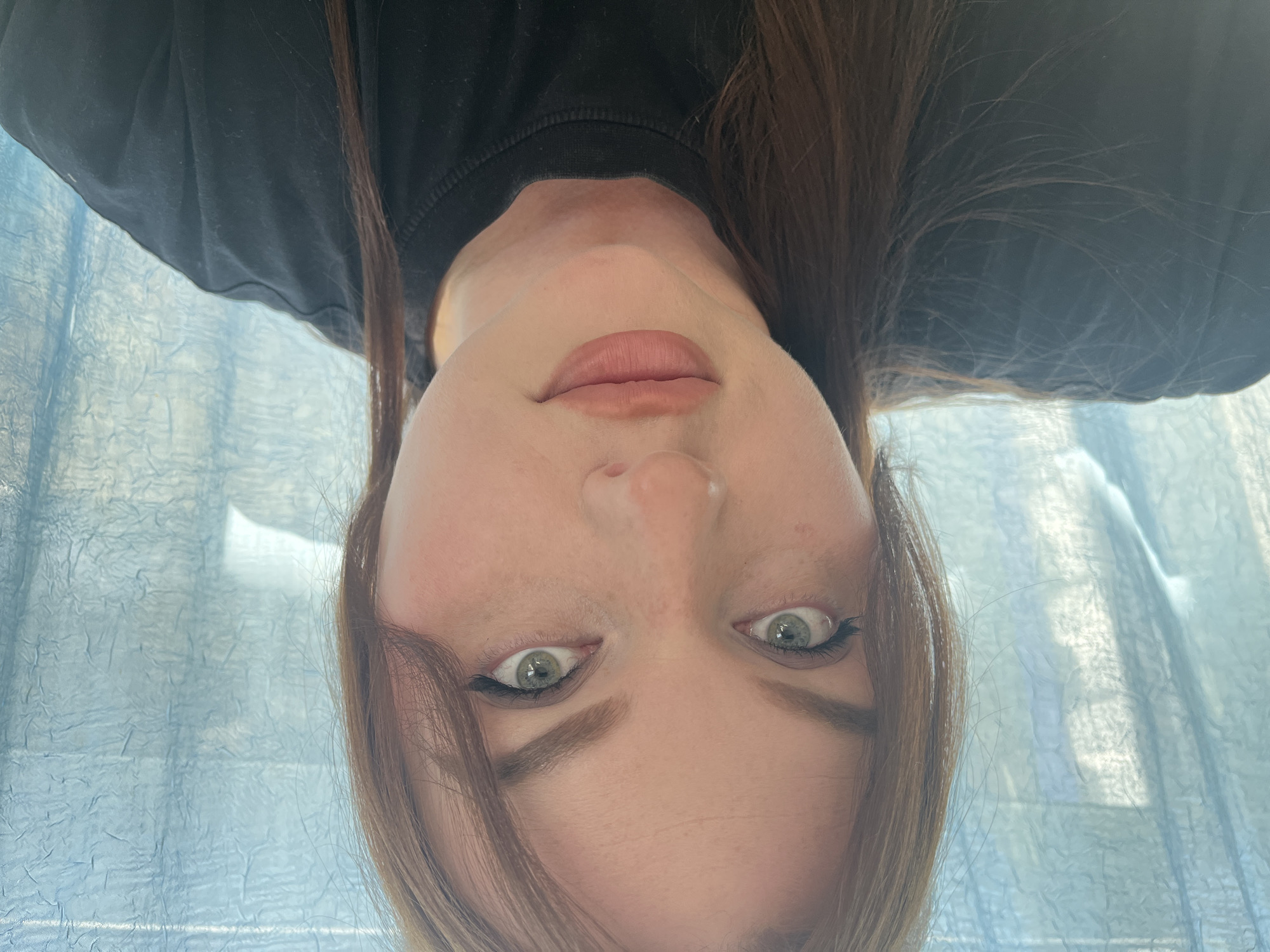 The image presents an inverted, close-up portrait of a person, likely female, captured from an unusual low-angle perspective. The subject has fair skin and striking green or hazel eyes, which are enhanced with dark eyeliner and mascara. Their eyebrows are well-defined and dark brown. Part of their face, from the lips upwards, is visible, with the lips showing a natural or light pink color. Long, dark brown hair frames the face, cascading from what would be the top of the head in a standard orientation. A dark-colored garment, appearing to be a black t-shirt or sweatshirt, is visible at the very top of the frame.
The background consists of a light blue, textured fabric, possibly a curtain or blind, which diffuses the light and creates a soft, almost ethereal backdrop. The lighting suggests it is daytime, with natural light filtering through the fabric. The person's expression is direct and neutral, looking straight into the camera with wide, attentive eyes. There is no discernible activity other than posing for the portrait. No visible text is present in the image. The setting appears to be indoors in Pavlovka, Russia.