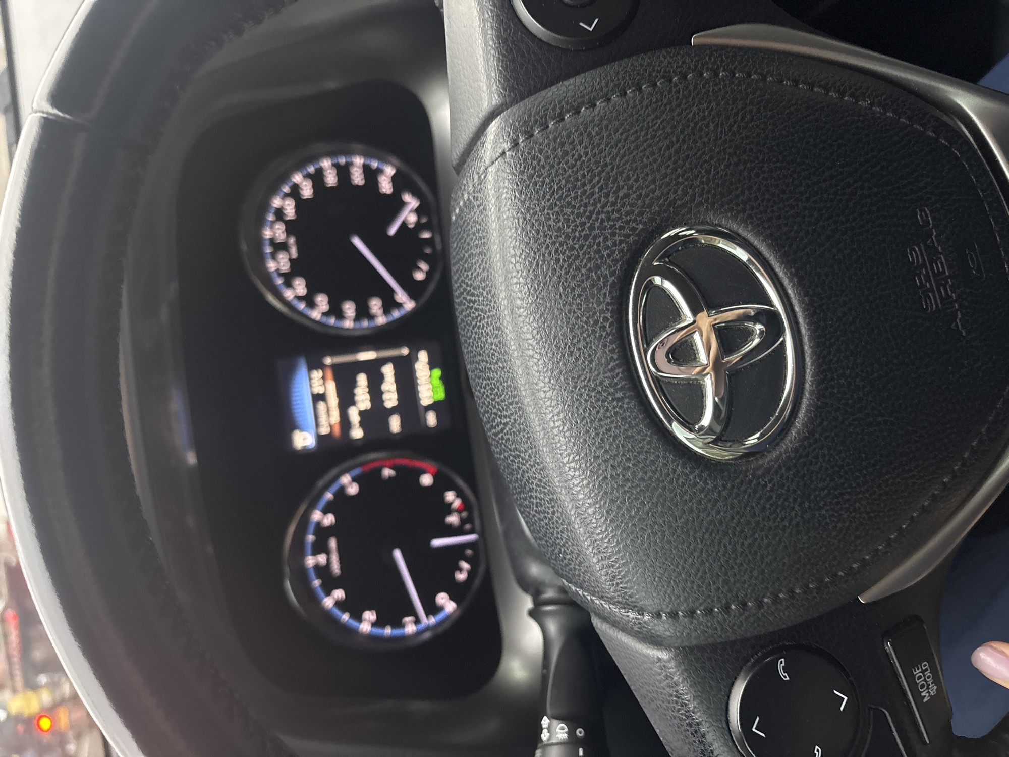 The image shows the steering wheel and instrument panel of a car, specifically a Toyota, based on the logo on the steering wheel. The instrument panel includes a speedometer and tachometer, with a digital display between them. The car's speed appears to be low, and the tachometer is at idle. The surrounding environment is not clearly visible, but suggests city driving, with the location determined to be Bello, Colombia. No people are fully visible, but a fingertip appears on the right side of the steering wheel.