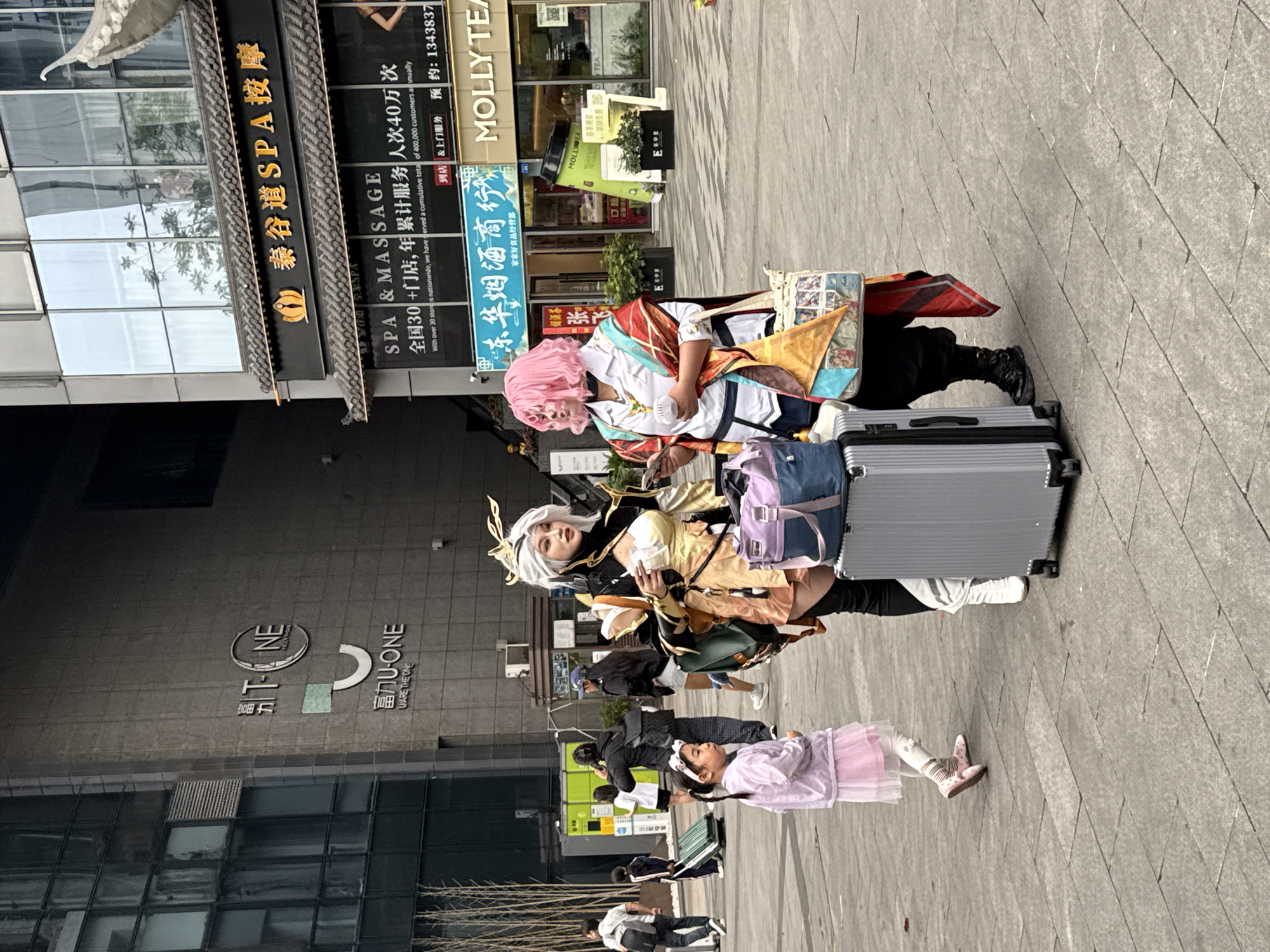 The image captures an urban street scene in Qingyang District, China, during the daytime. The main subjects are two individuals in elaborate cosplay costumes, standing with their luggage. One cosplayer wears a white, red, and gold outfit with pink hair, holding a small white fan and looking down at something in their hands. The other cosplayer wears a gold and black outfit with white/blonde hair and a headpiece, looking towards the viewer. They are positioned next to two suitcases, one silver and one dark, along with several bags including a backpack and tote bags.
In the foreground, a young girl in a pink dress and light-colored shoes is visible, looking to the left. In the background, other pedestrians are seen walking. The setting is a paved commercial area with modern buildings featuring large glass windows.
Visible text on the storefronts and buildings includes:
- "T-NE 富力U-ONE U ARE THE ONE" on a building facade.
- "泰谷道SPA按摩" (Thai Valley SPA & Massage), with an advertisement detailing "SPA & MASSAGE 全国30+门店,年累计服务人次40万次 With over 30 stores nationwide, we have served a cumulative total of 400,000 customers annually 到店&上门服务 预约:1343837".
- "东华烟酒商行" (Donghua Tobacco & Alcohol Shop).
- "MOLLY TEA" appearing twice, with additional text like "春茶观音" (Spring Tea Guanyin) and "波排生香 TEA".
- Other smaller texts like "张飞" and "家家好食品经营部".
The bright lighting suggests clear weather. The cosplayers appear to be paused, possibly resting or posing, amidst the bustle of the street.
