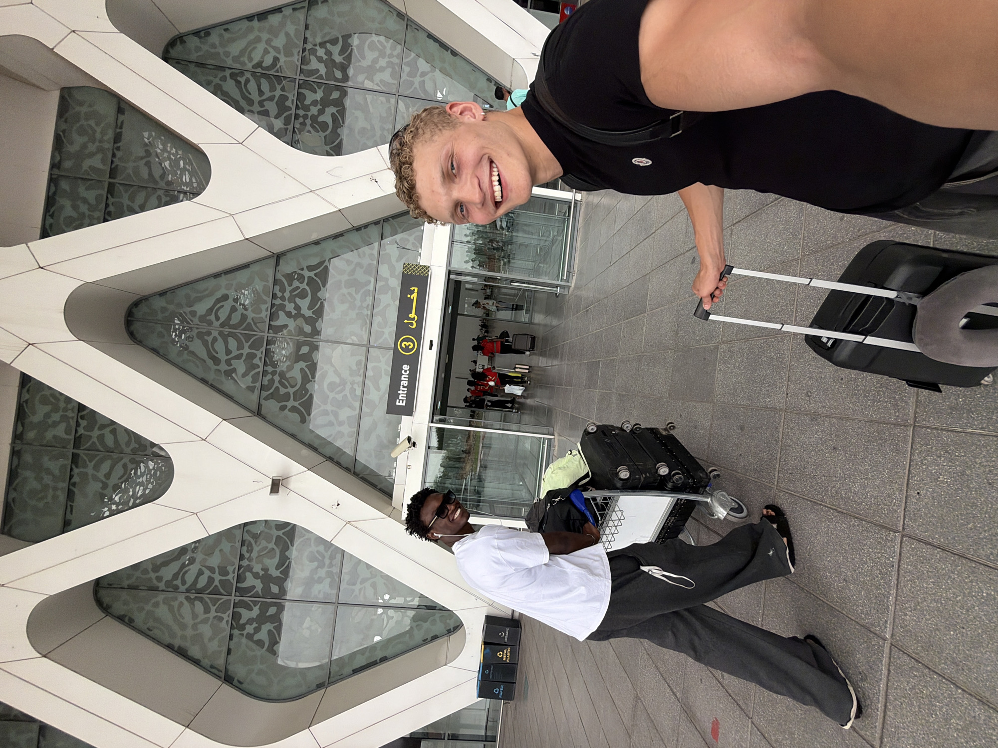 The image depicts two young men at Marrakesh Menara Airport (RAK), Morocco. The airport's distinctive modern architecture with geometric patterns on the facade is visible in the background.
The man on the left, with dark skin and braided hair, is wearing sunglasses, a white t-shirt, and dark loose-fitting pants. He is pushing a luggage cart filled with bags. He is smiling and looking towards the camera.
The man on the right, with lighter skin and curly blond hair, is wearing a black t-shirt and has a backpack on. He is holding the handle of a black rolling suitcase and is smiling broadly as he takes a selfie.
The entrance to the airport is visible behind them, with signage including "Entrance 3" and Arabic text. Several other people are seen entering and exiting the airport. Recycling bins are also visible to the left of the entrance. The overall scene suggests the beginning or end of a trip.