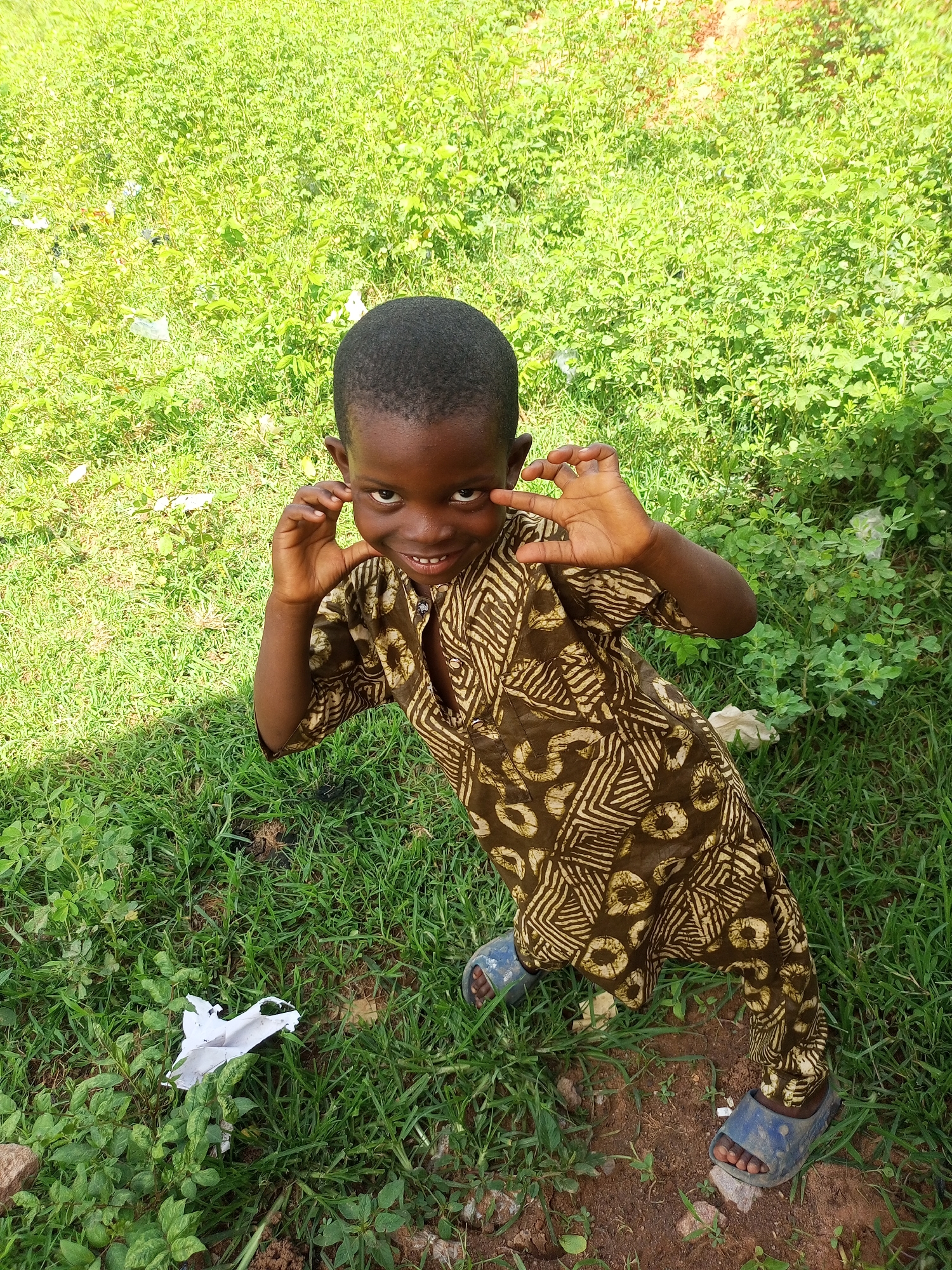 In the city of Tempure, Nigeria, a young Black boy, estimated to be between four and six years old, stands outdoors on a patch of green grass. He is wearing a loose-fitting, long-sleeved tunic with a brown and tan geometric pattern, and blue sandals. The boy is looking directly at the camera with a playful expression, holding his hands up to his face as if playing peek-a-boo. Sunlight casts shadows on the ground, indicating it is daytime. The background is a blur of green vegetation and some scattered white paper debris.