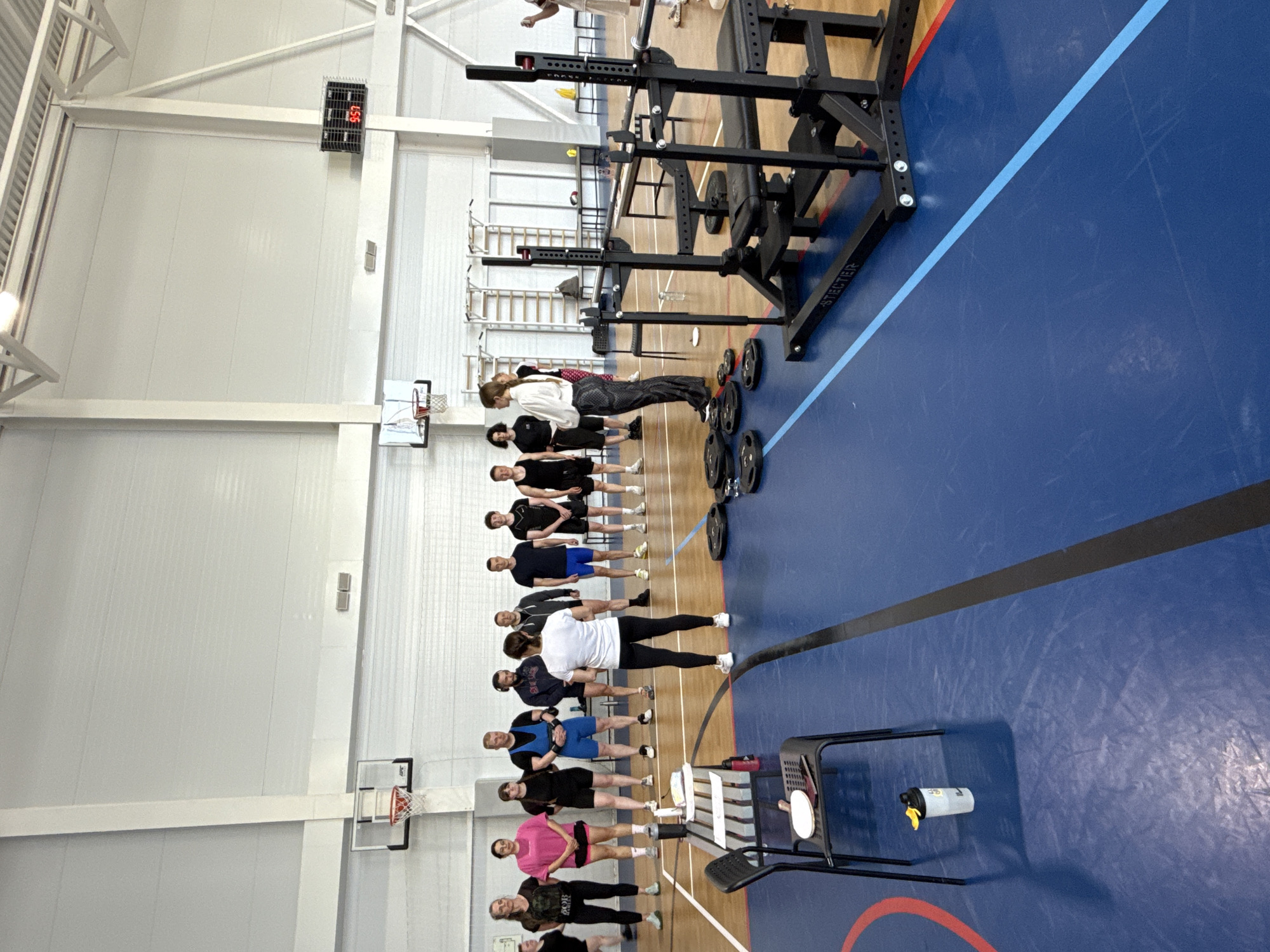 The image captures a training scene in what appears to be an indoor sports hall, potentially located in Votkinsk, Russia. A group of approximately 15 people is lined up, facing the same direction. Most individuals wear athletic attire, suggesting participation in a workout or training session. In the foreground, a weight training rack and weight plates are visible, while a drinking bottle and a black chair are in the lower-right corner. The background features a basketball court and a scoreboard showing '4:57'. The hall has white walls and a high ceiling with visible supporting beams. The floor is a mix of blue, brown, and red zones.