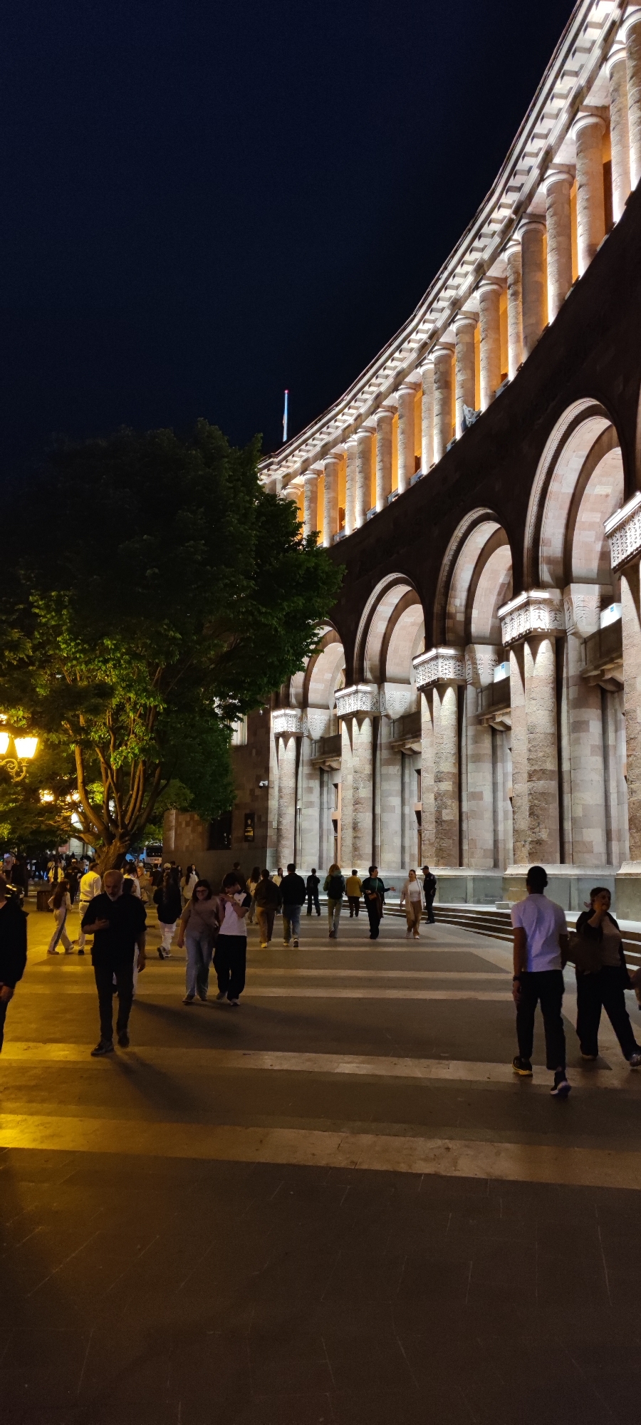 At night, a crowded sidewalk in Yerevan, Armenia, is illuminated by street lamps and lights from a large building. The building features multiple arches and columns, with lights shining from within each arch. A flag is visible atop the building. Pedestrians, a mix of men and women, walk along the sidewalk. Some are walking in groups, while others are alone. The sidewalk is dark and marked with light-colored stripes. A large tree with green leaves stands near the sidewalk.