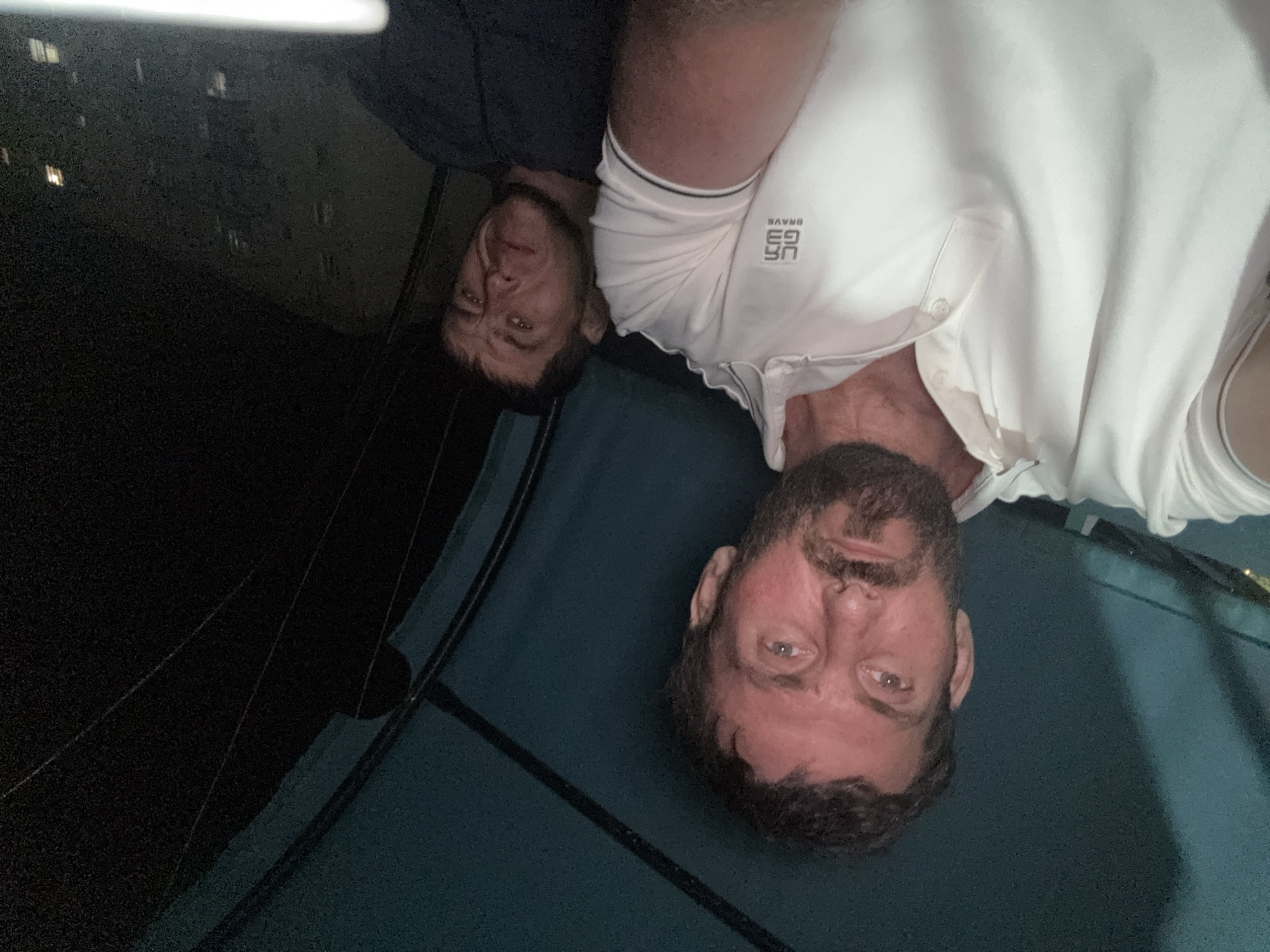 A selfie taken at night shows two men sitting close together under a dark canopy. The man in the foreground, on the left, is wearing a white polo shirt and has a beard. He is looking directly at the camera. The man in the background, on the right, is wearing a dark t-shirt and has a concerned expression. He is looking slightly off-camera. The background is dark, with the silhouette of a building visible on the right side of the image. The overall lighting suggests it is nighttime.
